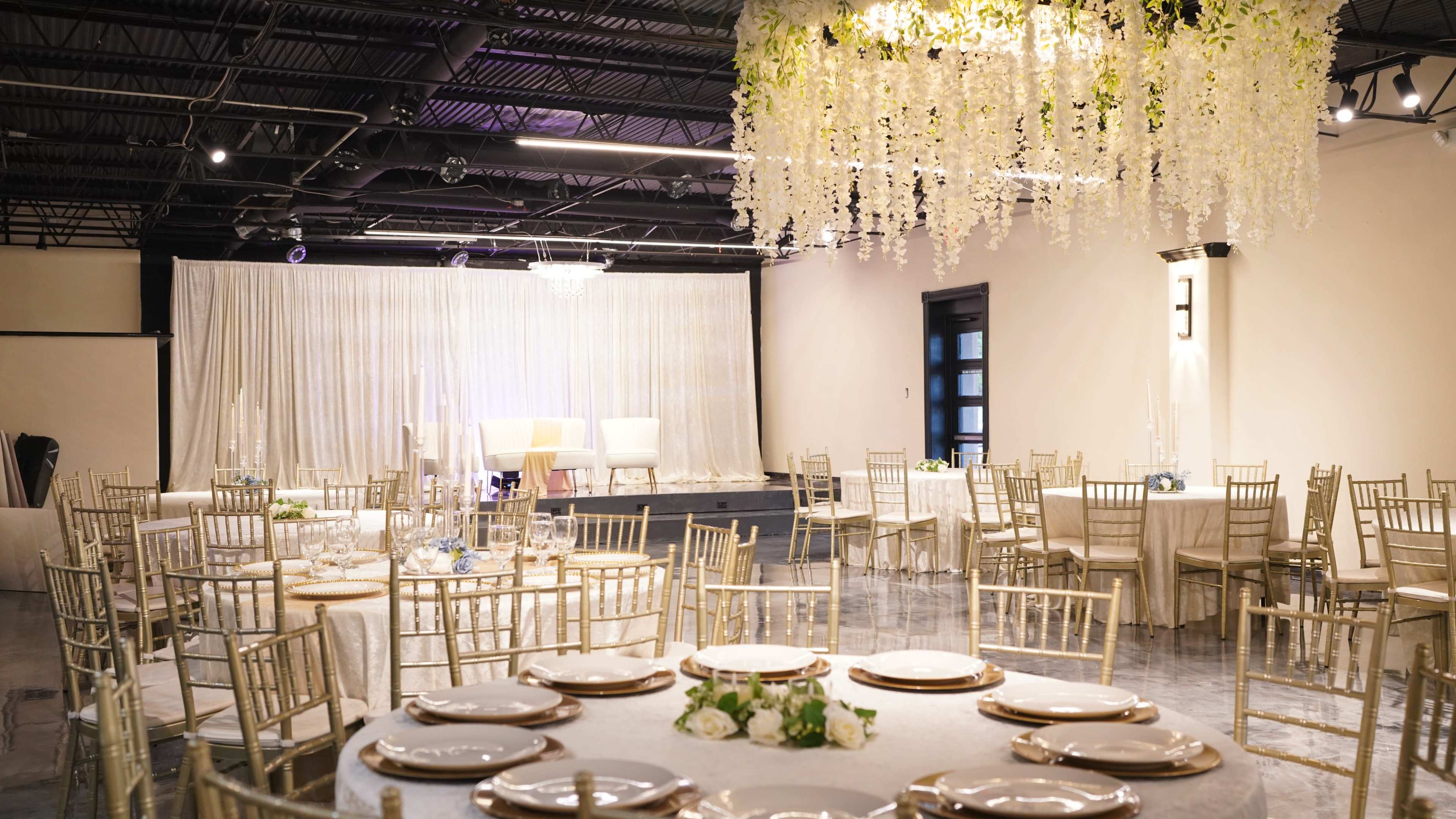 The image shows a banquet hall set for a formal event, featuring round tables with gold chairs, elegant centerpieces, and a stage adorned with drapery and a chandelier.