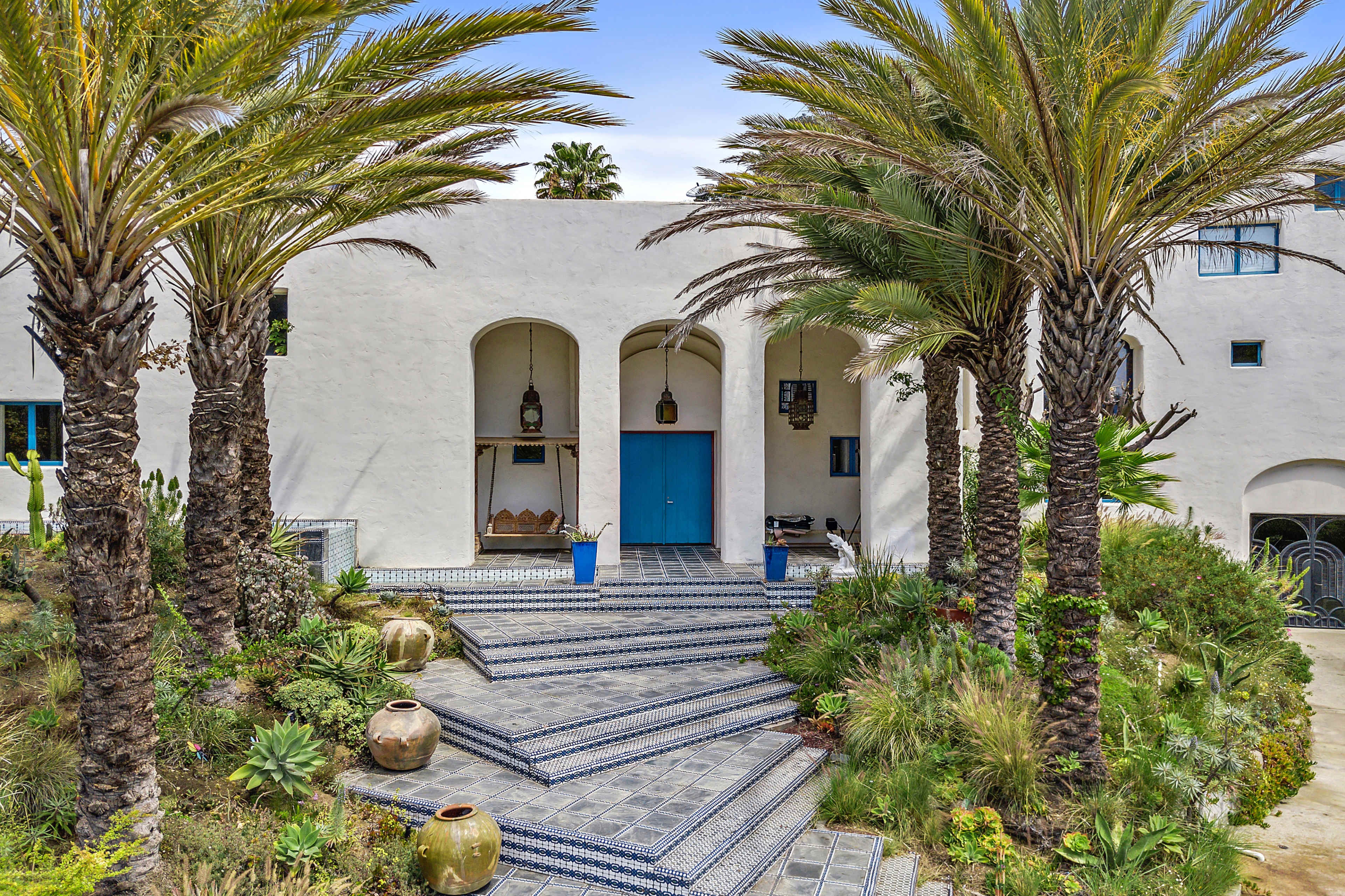 The image shows a Mediterranean-style home with a blue door, flanked by palm trees and a tiled staircase leading to the entrance.