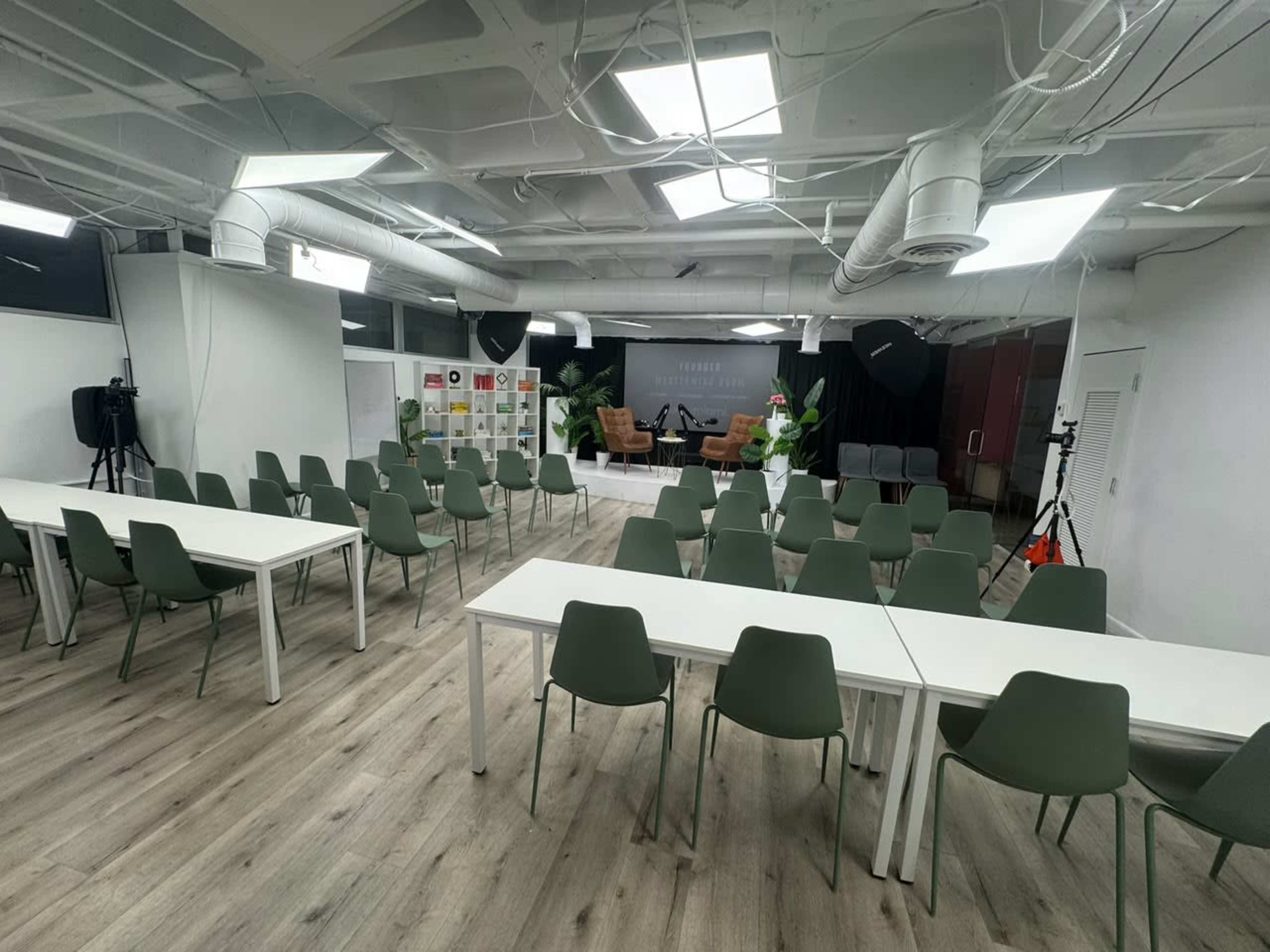 A spacious event room is set up with several tables and chairs facing a small stage, which features seating and a backdrop.