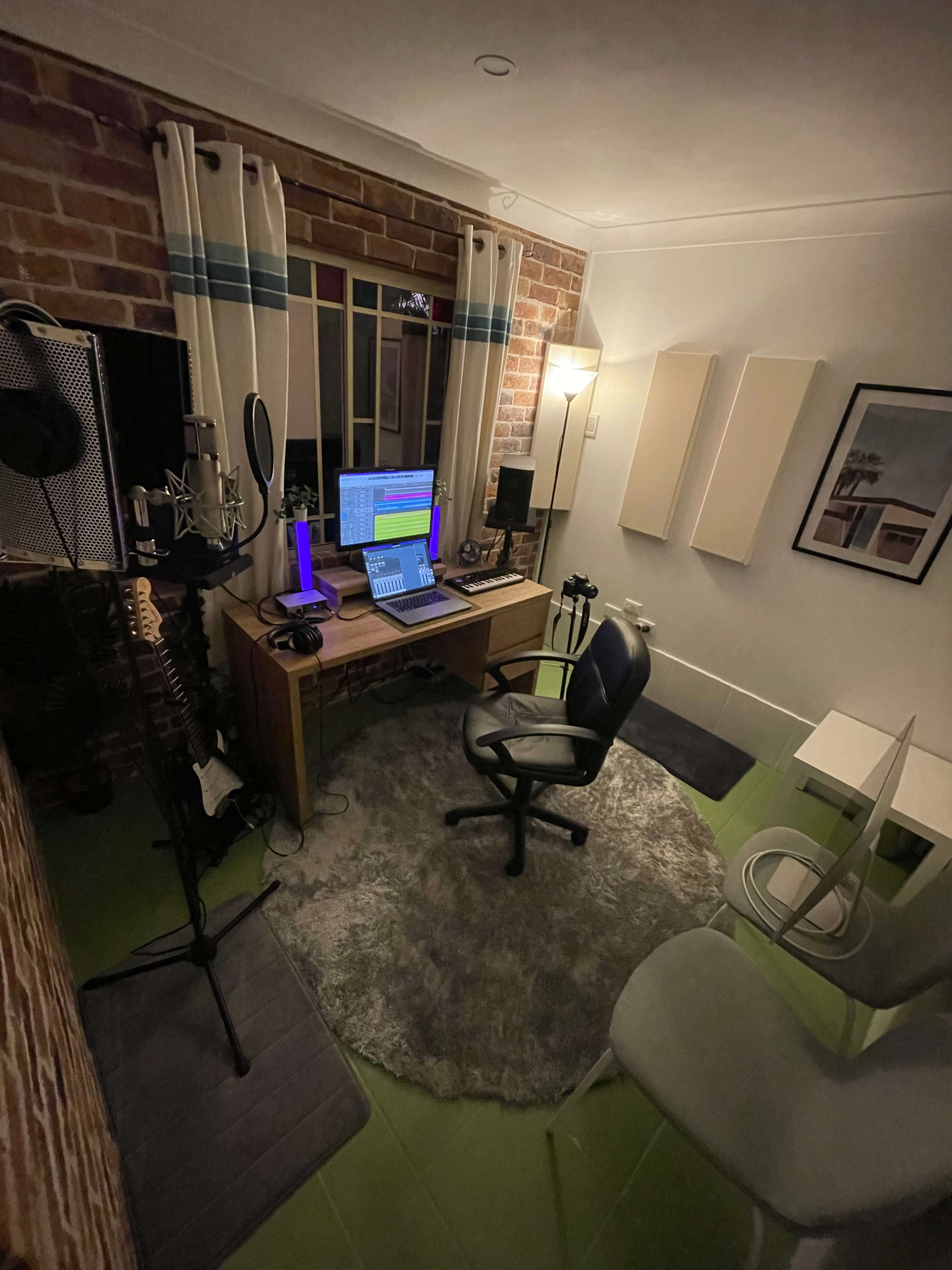 A small music production studio featuring a desk with a computer, a microphone setup, and various instruments arranged around a rug.