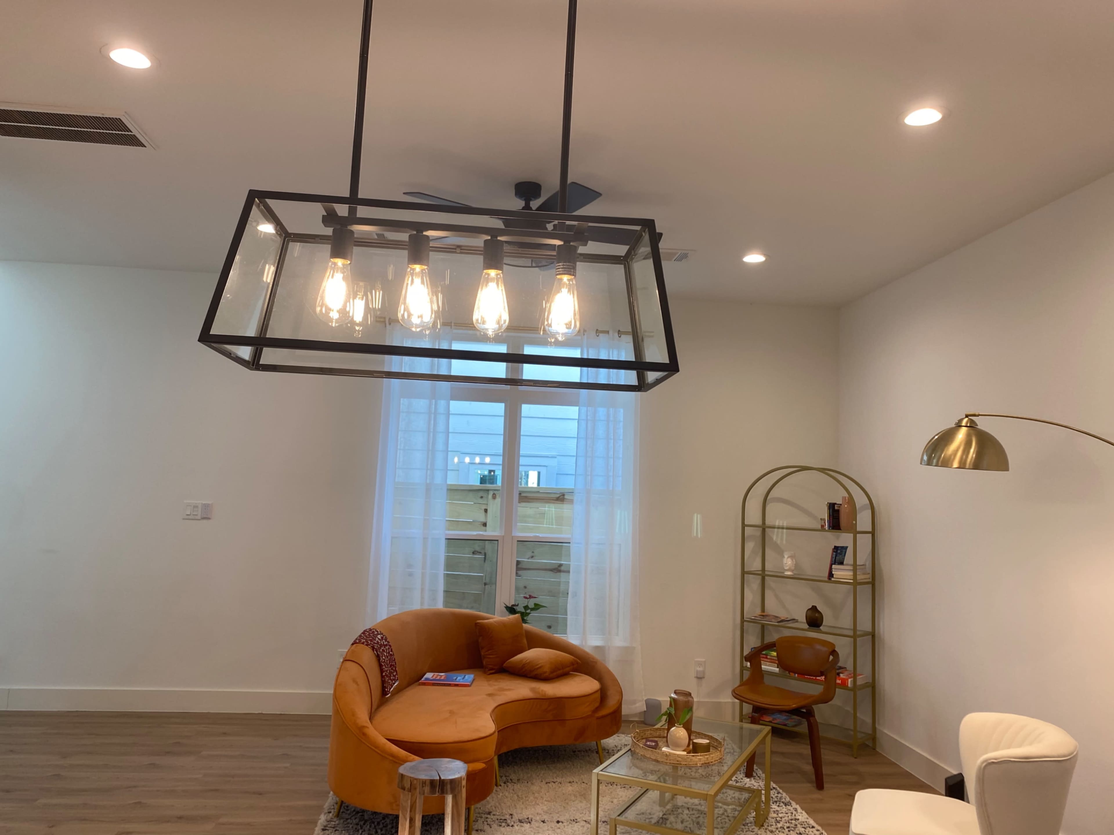 A modern living room with a curved orange sofa, a glass coffee table, and a decorative shelving unit beneath a large, hanging light fixture.