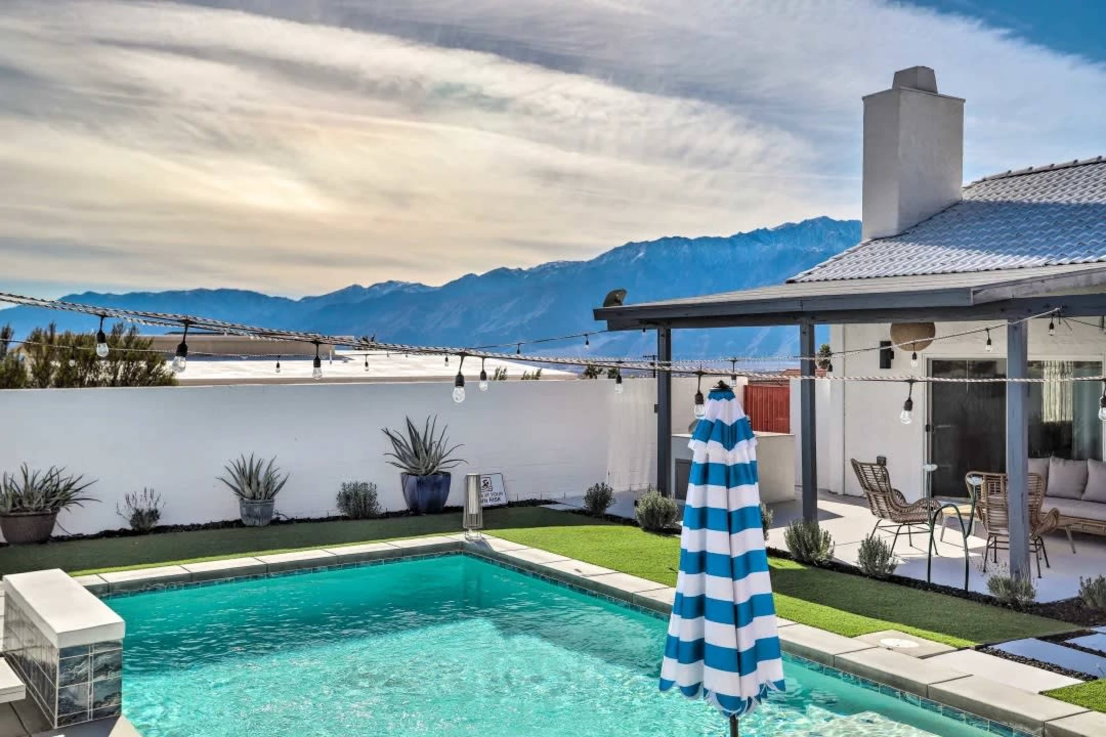 A swimming pool with a striped umbrella is situated beside a house, framed by mountains in the background.