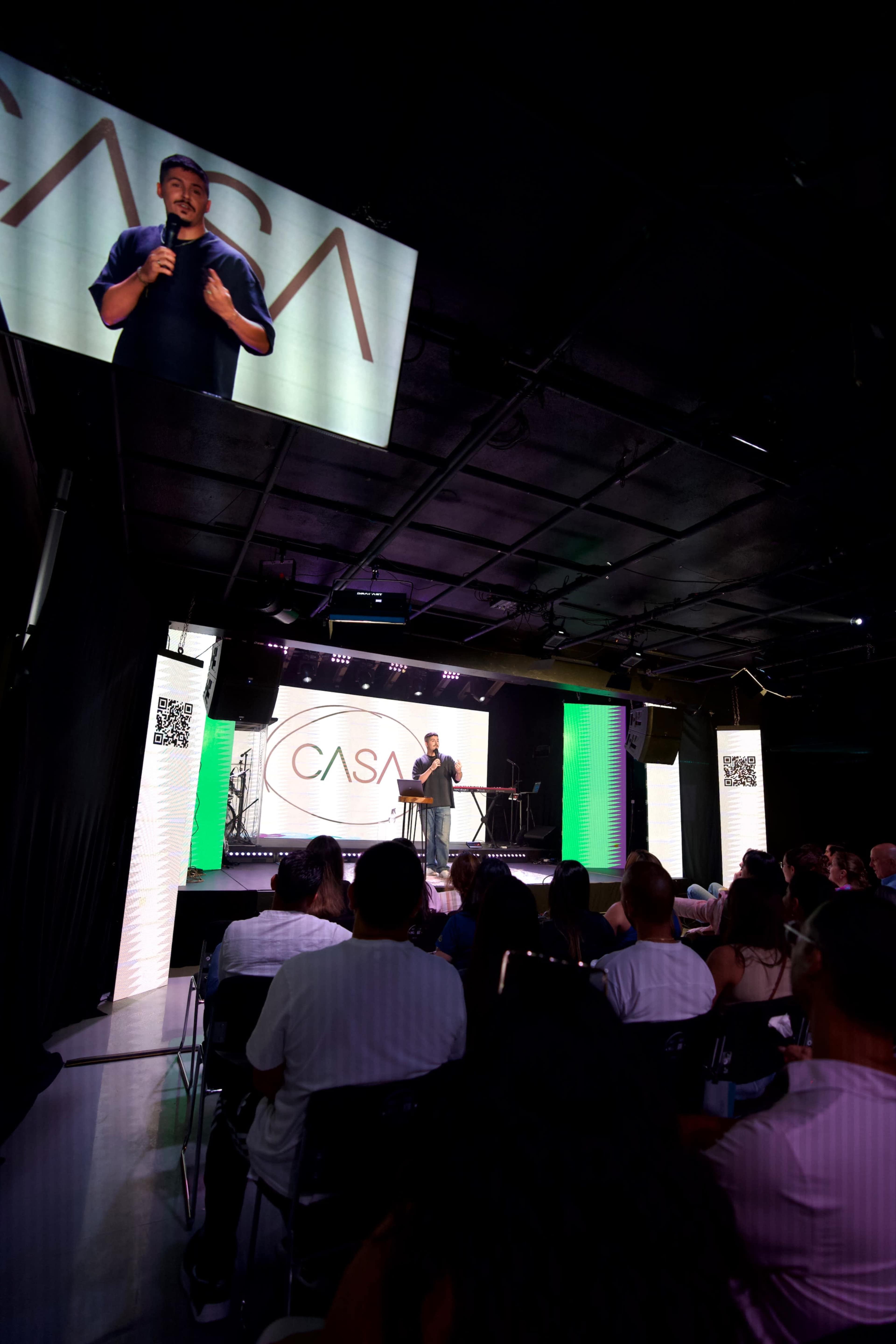 A speaker stands on stage in front of an audience, with a large screen displaying the word "CASA" behind them.