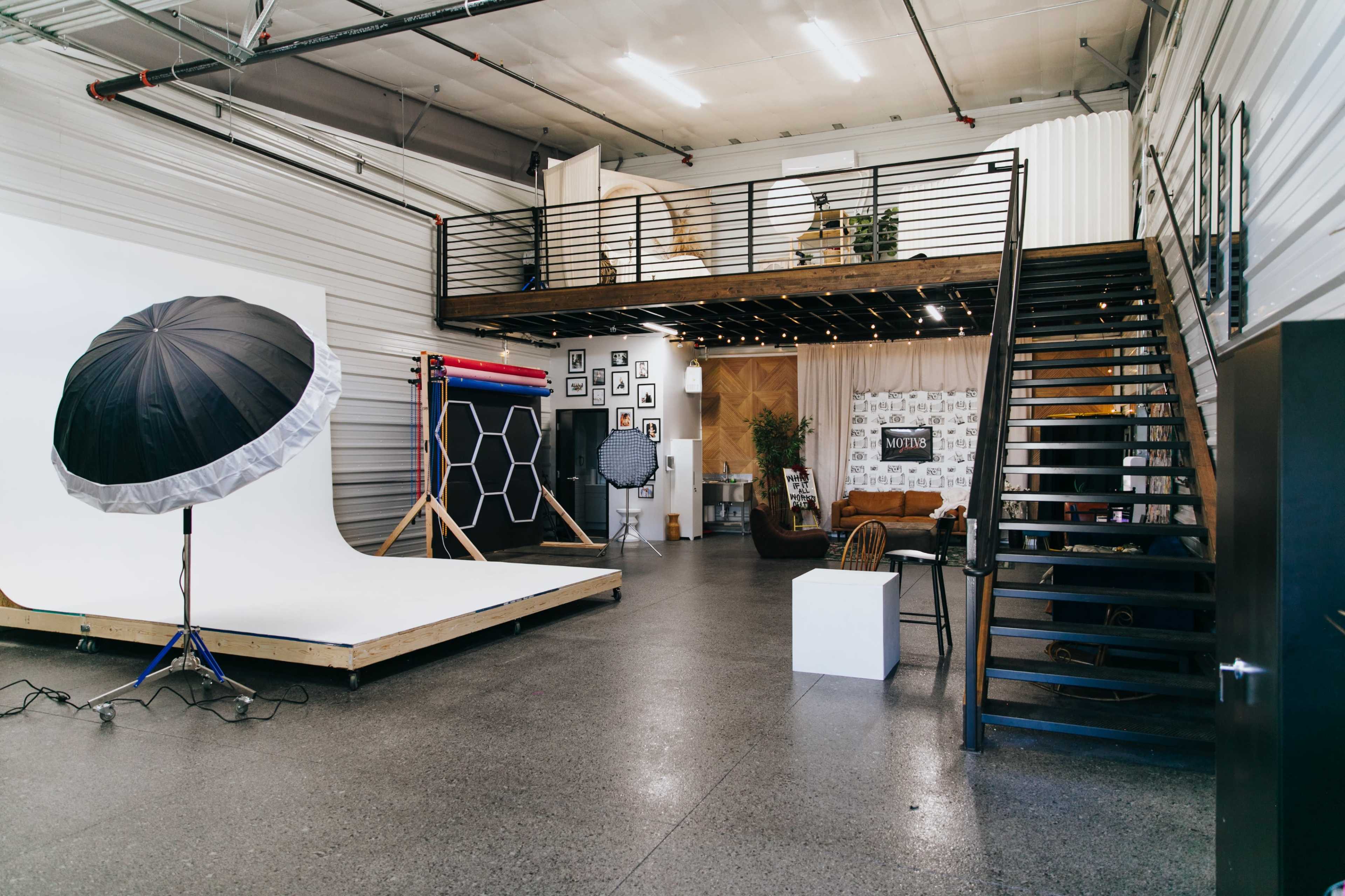 The image shows a spacious photography studio with a raised loft, a large backdrop area, and various photo equipment arranged throughout.