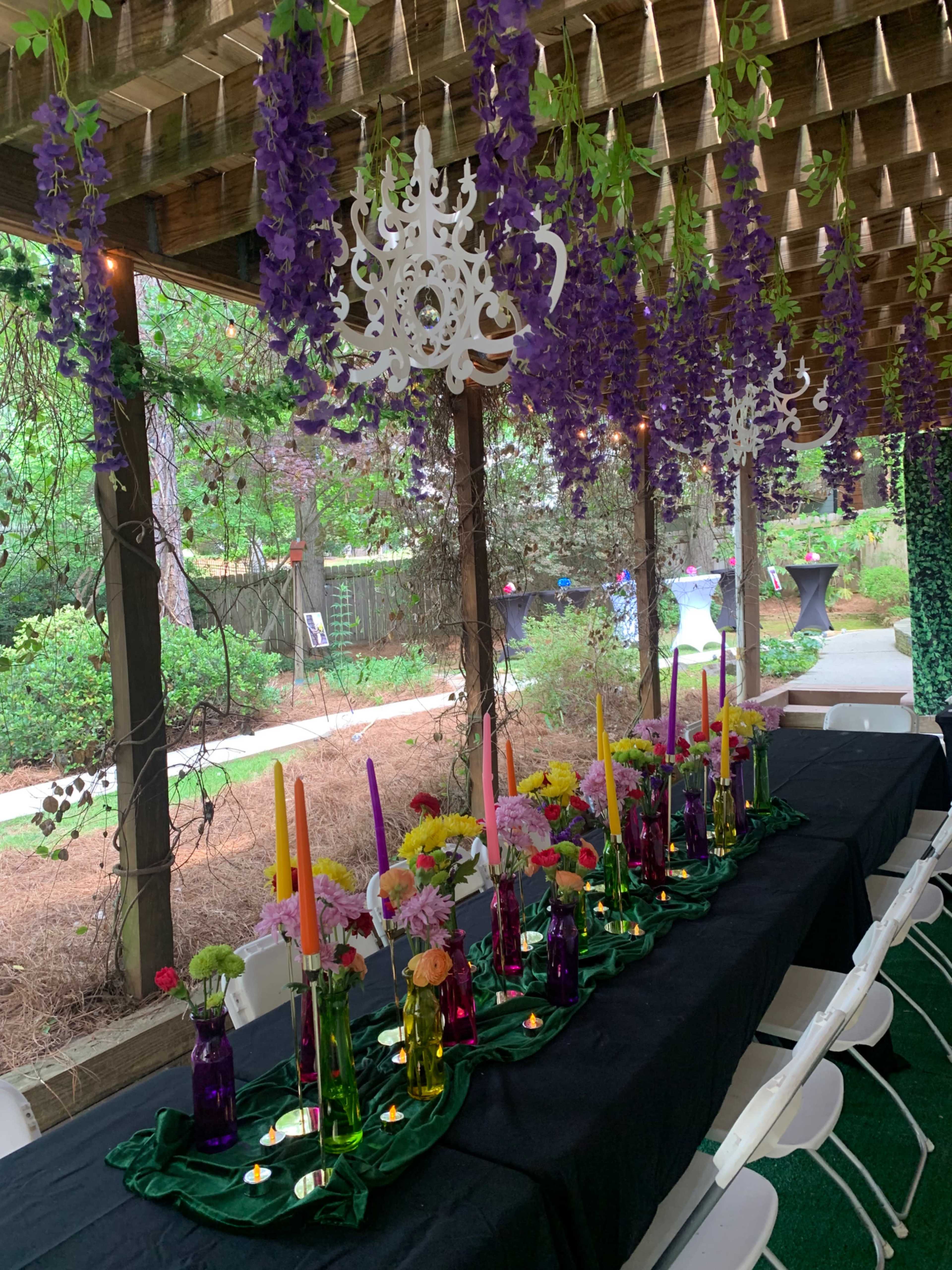 A long table adorned with colorful candles and floral arrangements is set beneath a wooden pergola draped in greenery and hanging decorations.