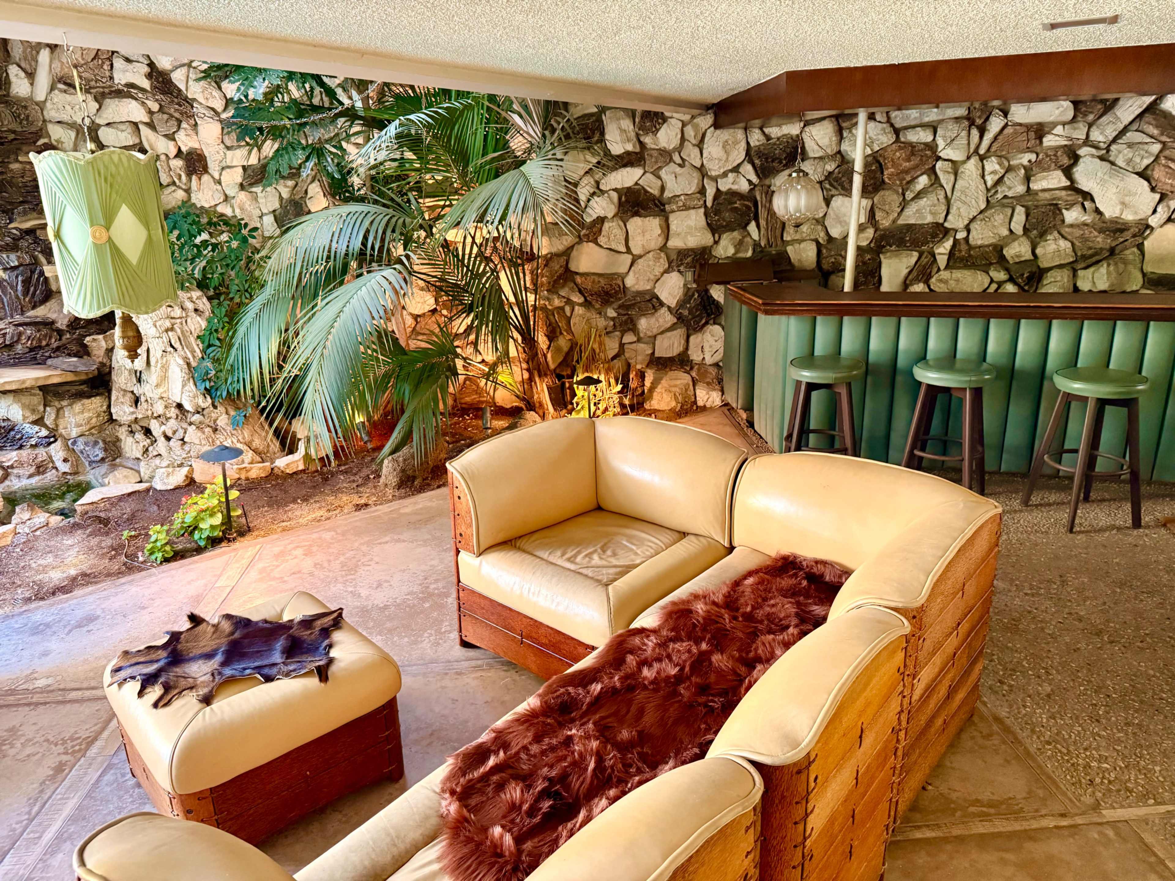 The image depicts a mid-century modern living room featuring a leather couch, a fur throw, a stone wall backdrop, and a small bar area with green seating.