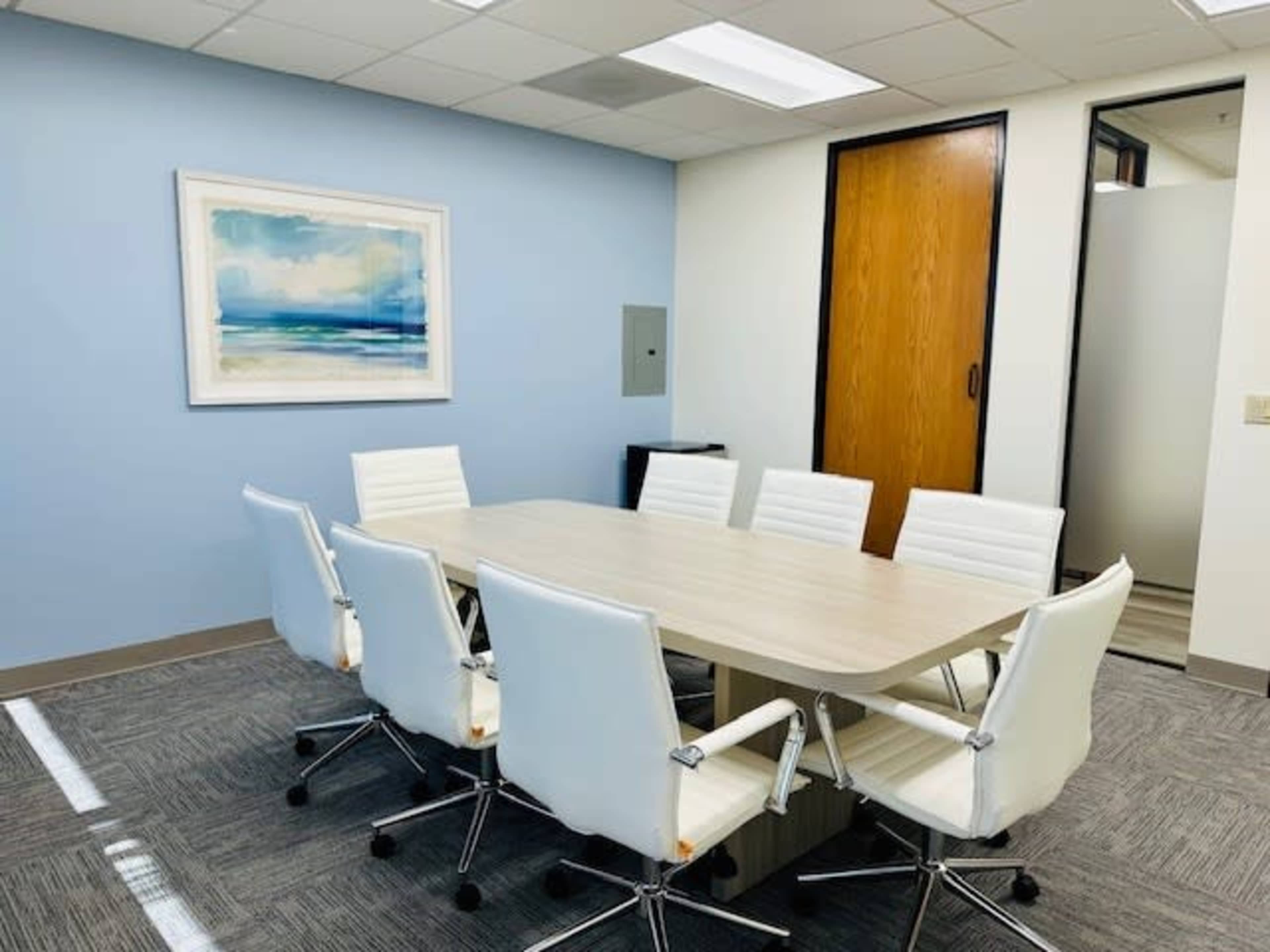 The image shows a conference room with a rectangular table surrounded by white chairs, a blue wall, and a framed piece of artwork.