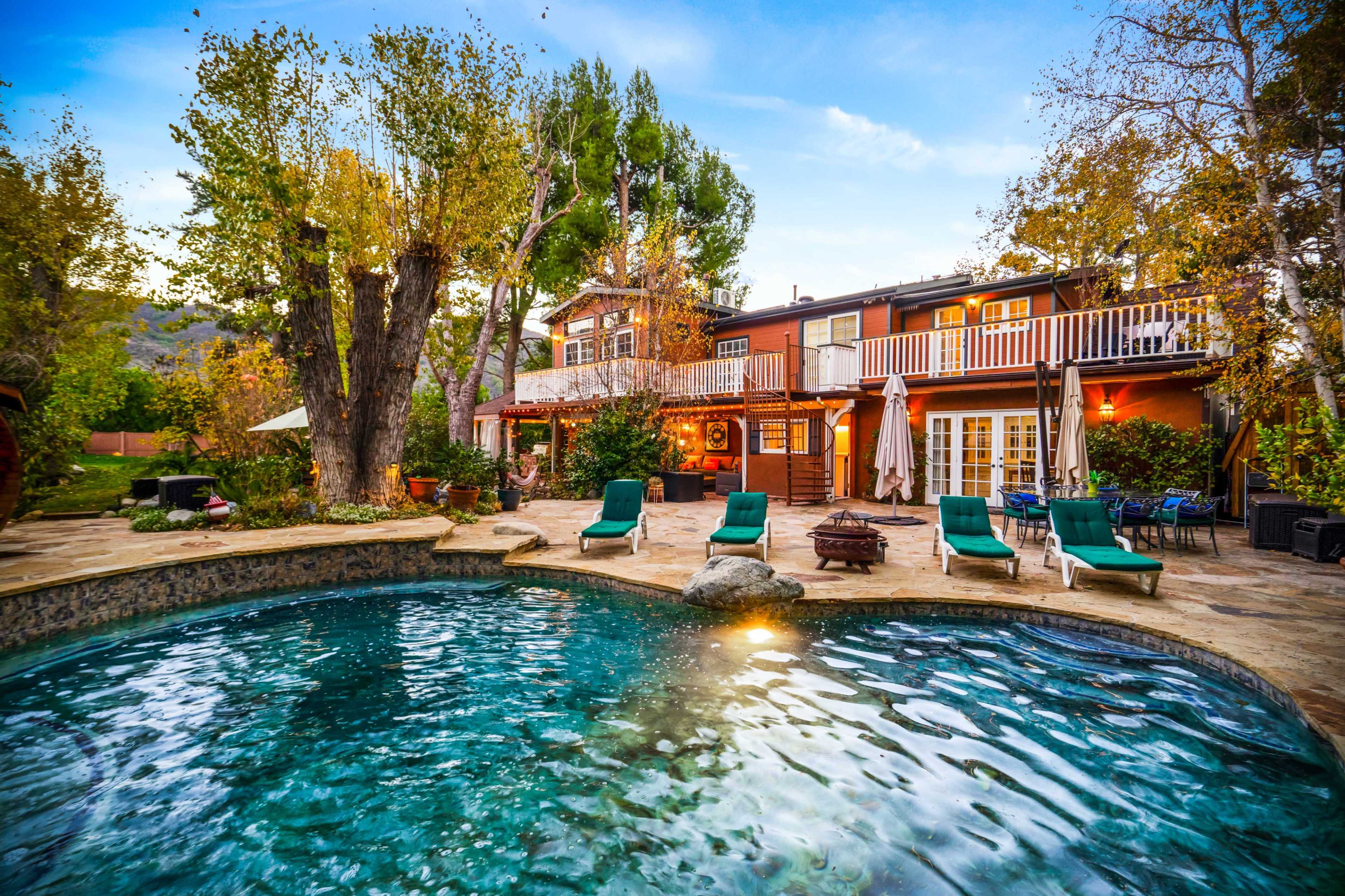The image shows a backyard with a swimming pool, lounge chairs, and a two-story house surrounded by trees.