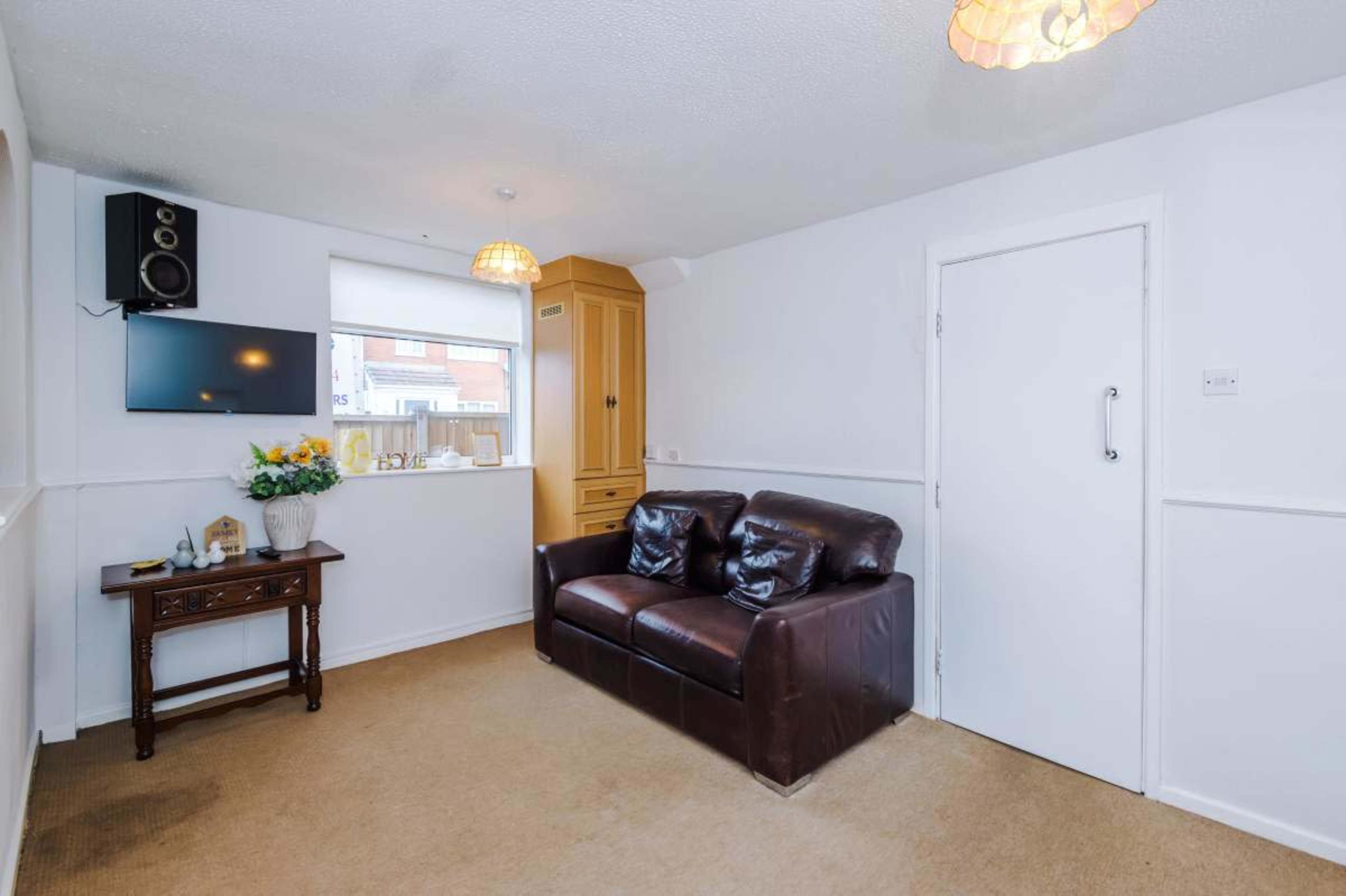 The image shows a small living room with a brown leather sofa, a wall-mounted television, and a wooden side table adorned with decorative items.