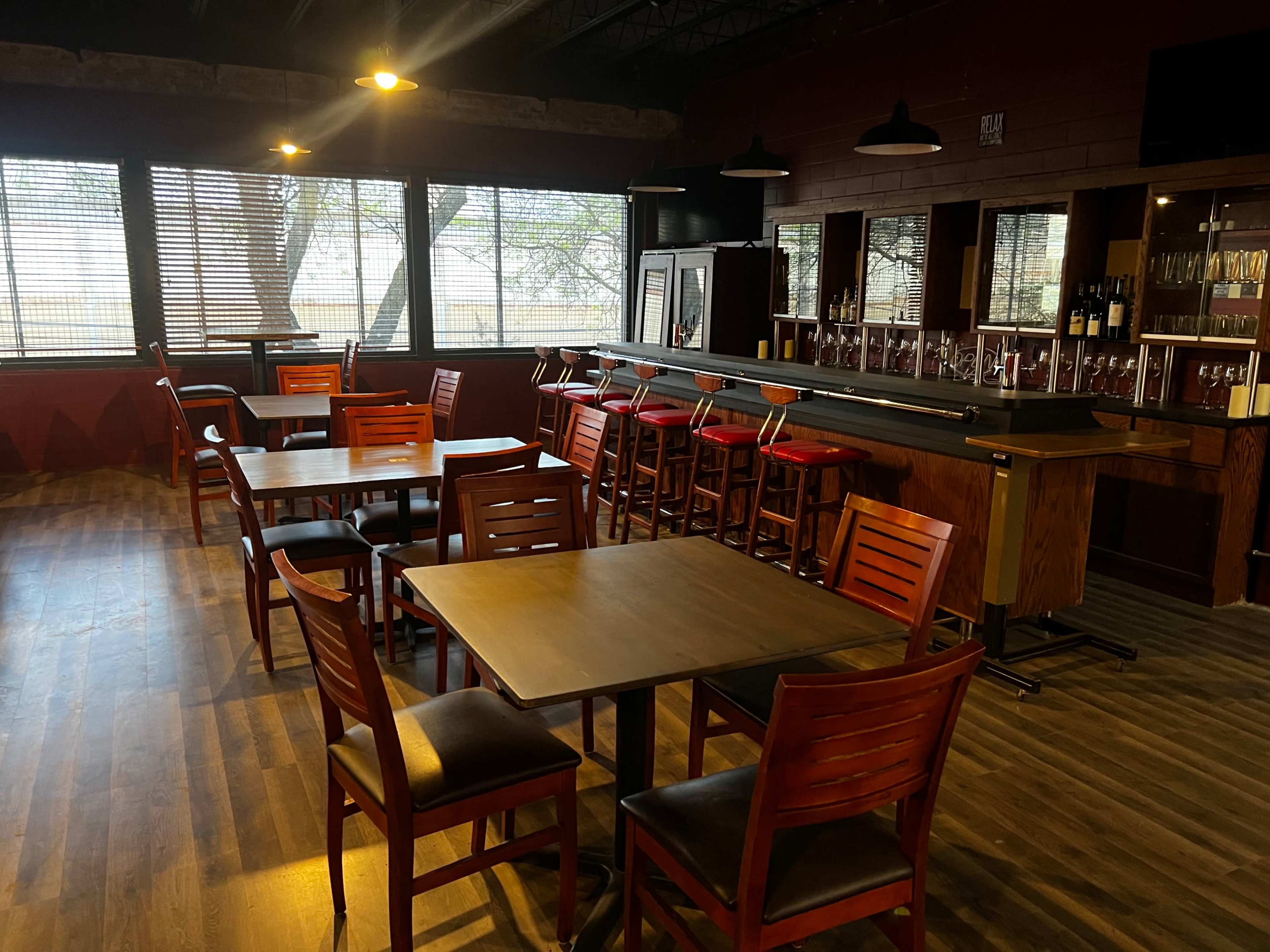 An empty bar area with wooden tables and chairs, a counter with bar stools, and large windows letting in natural light.