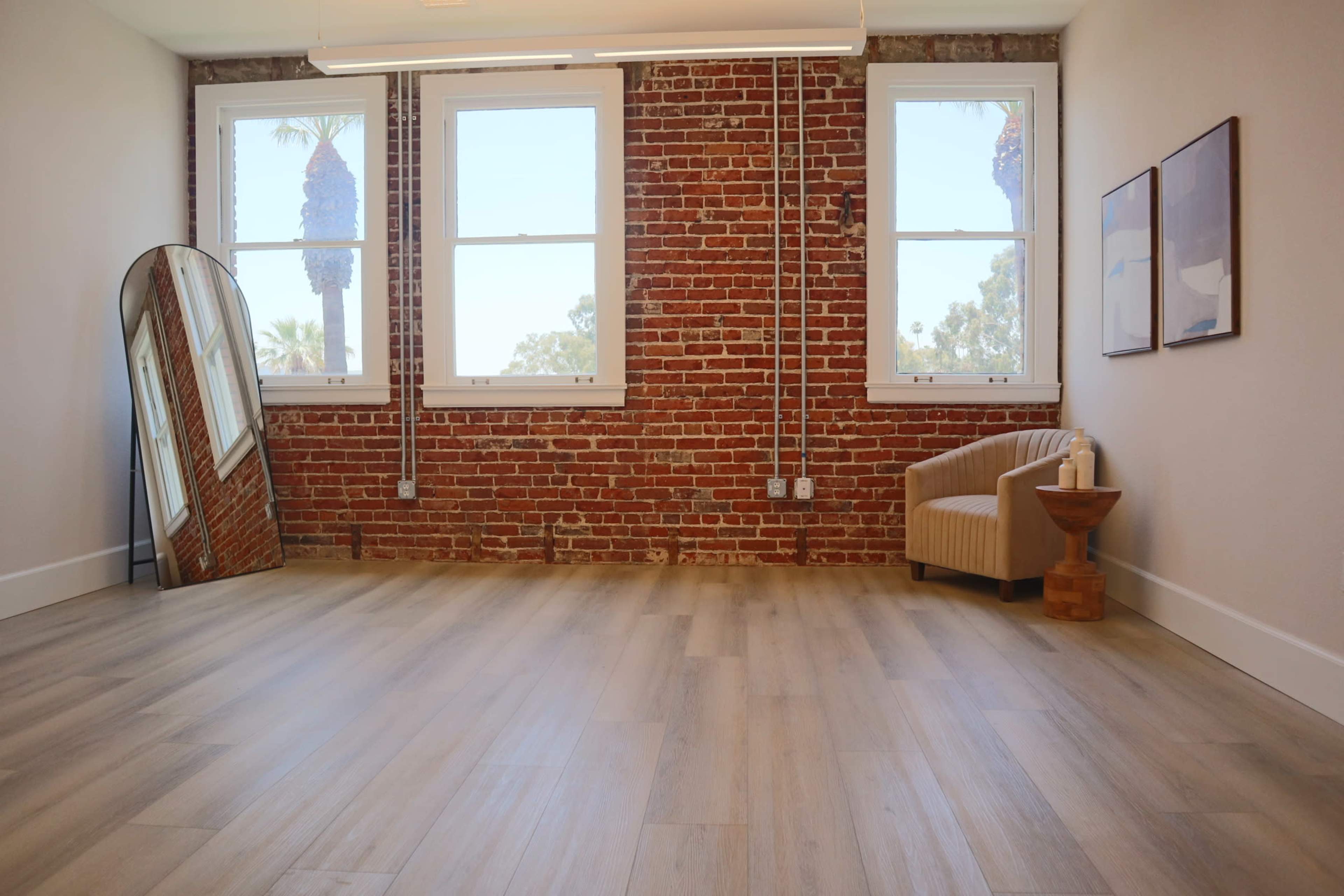 A spacious room with a brick wall, large windows, a curved mirror, a small wooden side table, and an upholstered chair.