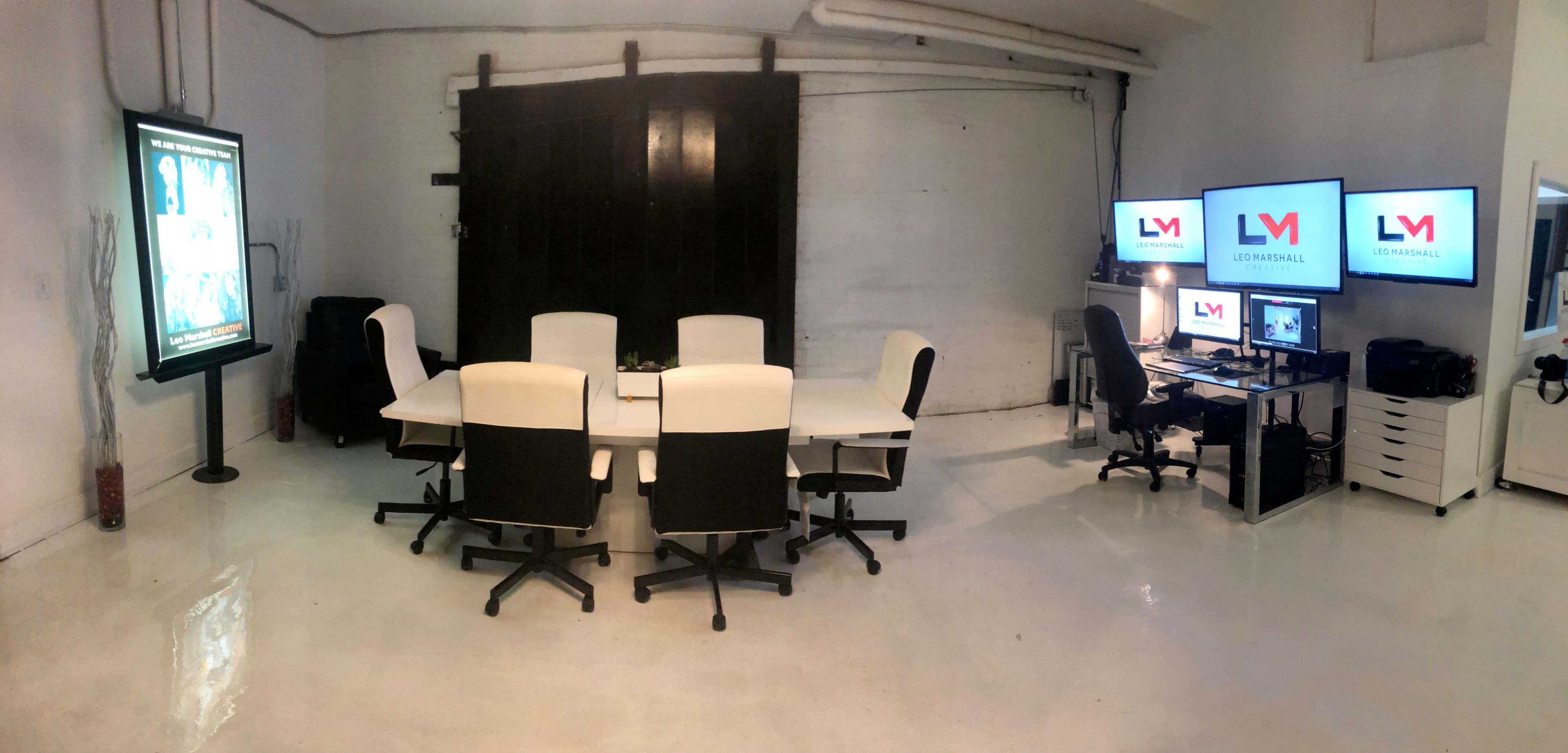 A modern office space featuring a large conference table with six chairs, two computer monitors, and a display screen on one wall.