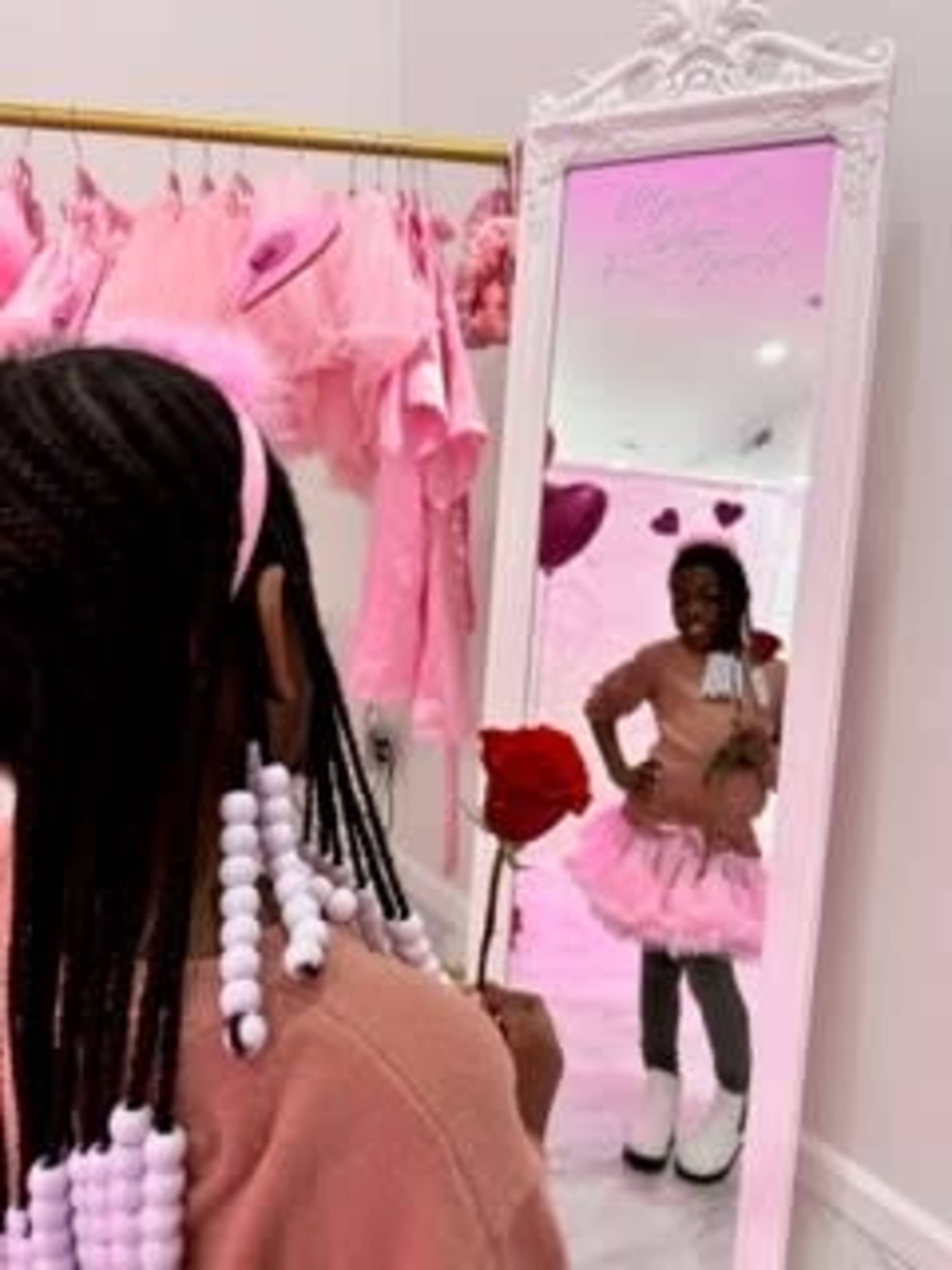 A child in a pink outfit holds a rose while posing in front of a mirror surrounded by pink clothing and decorations.