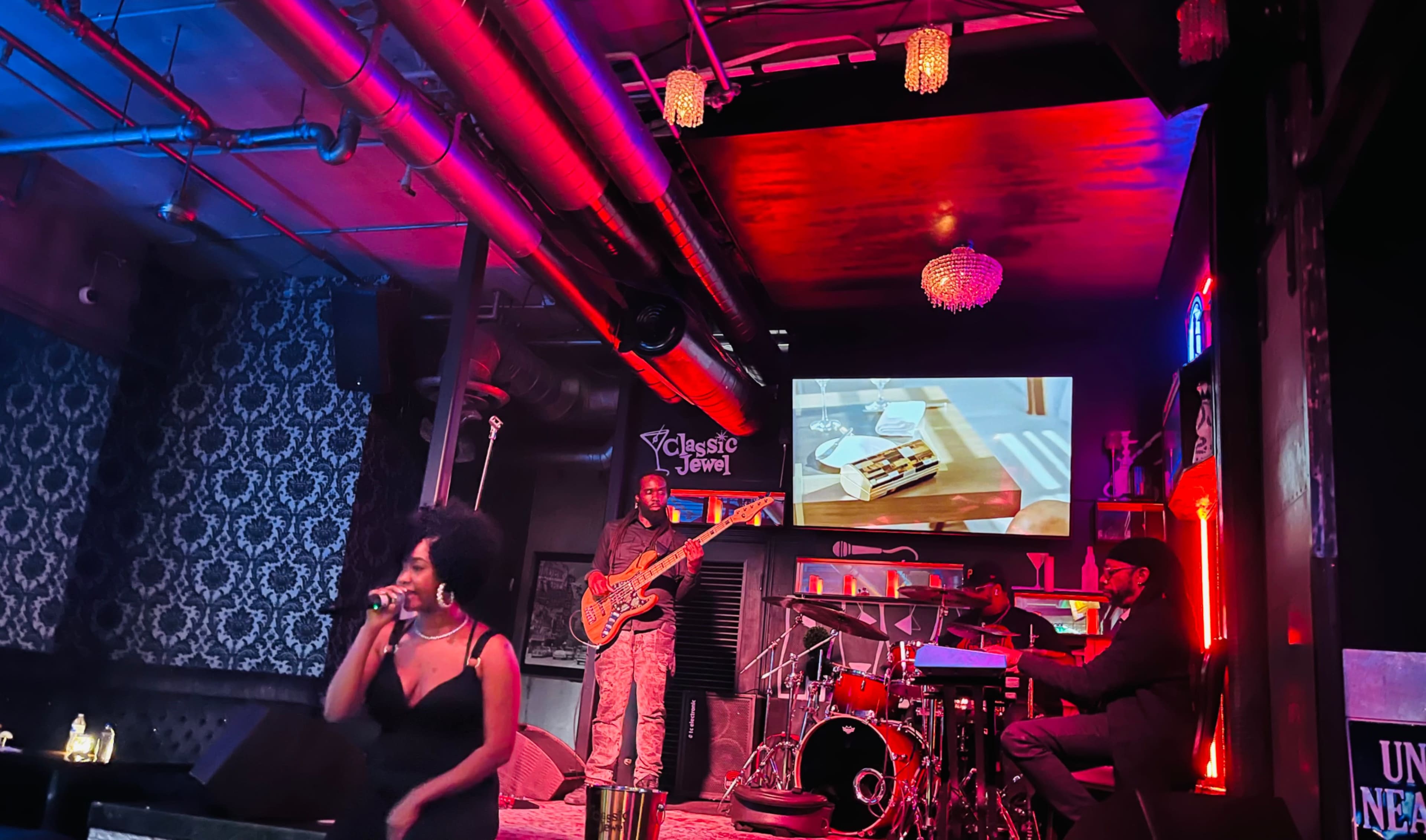 A woman sings on stage in front of a band while a video plays on a screen behind them in a dimly lit venue.