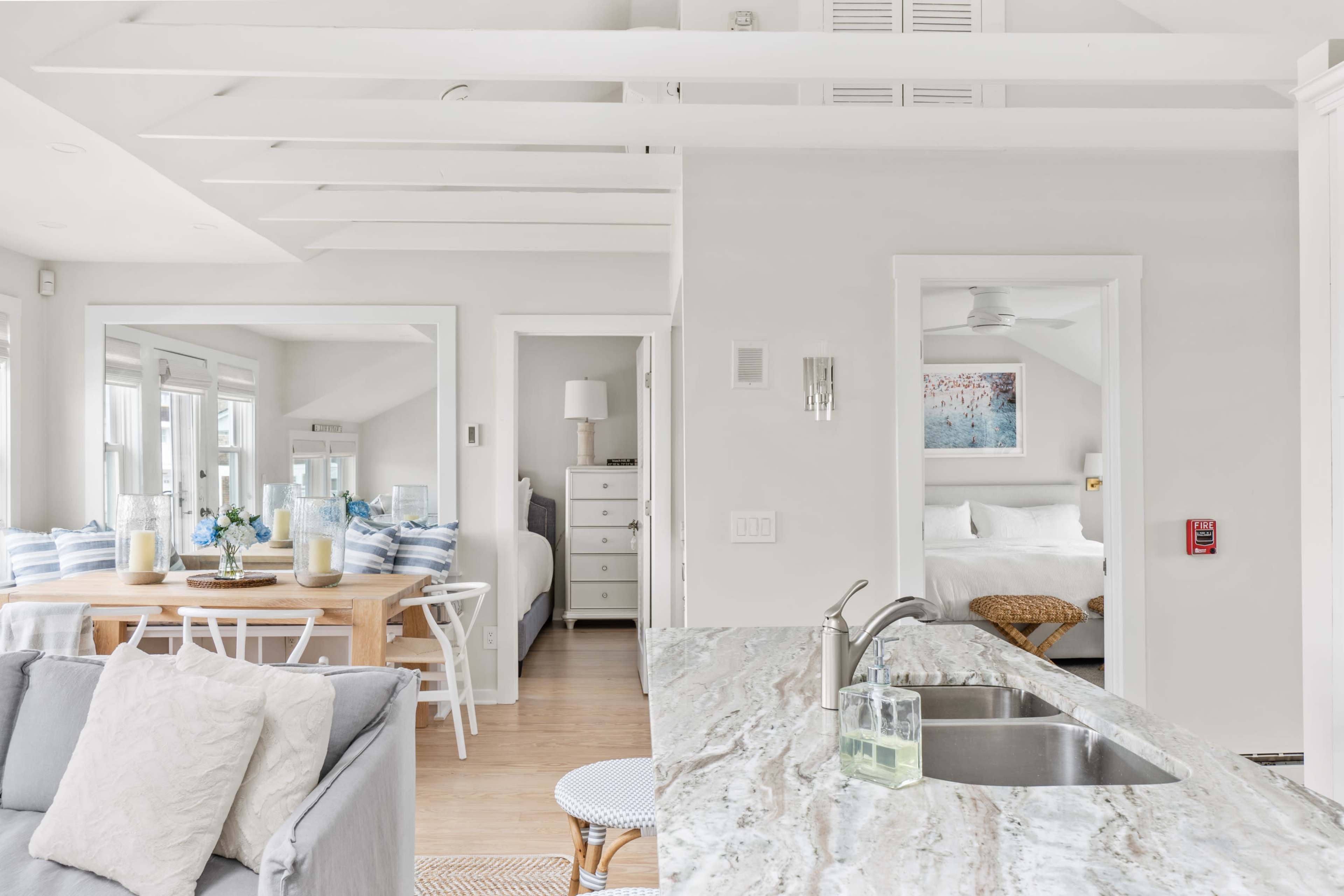 A bright, open-concept living space features a dining area with a wooden table, a kitchen with a marble countertop, and a bedroom visible through an adjoining door.