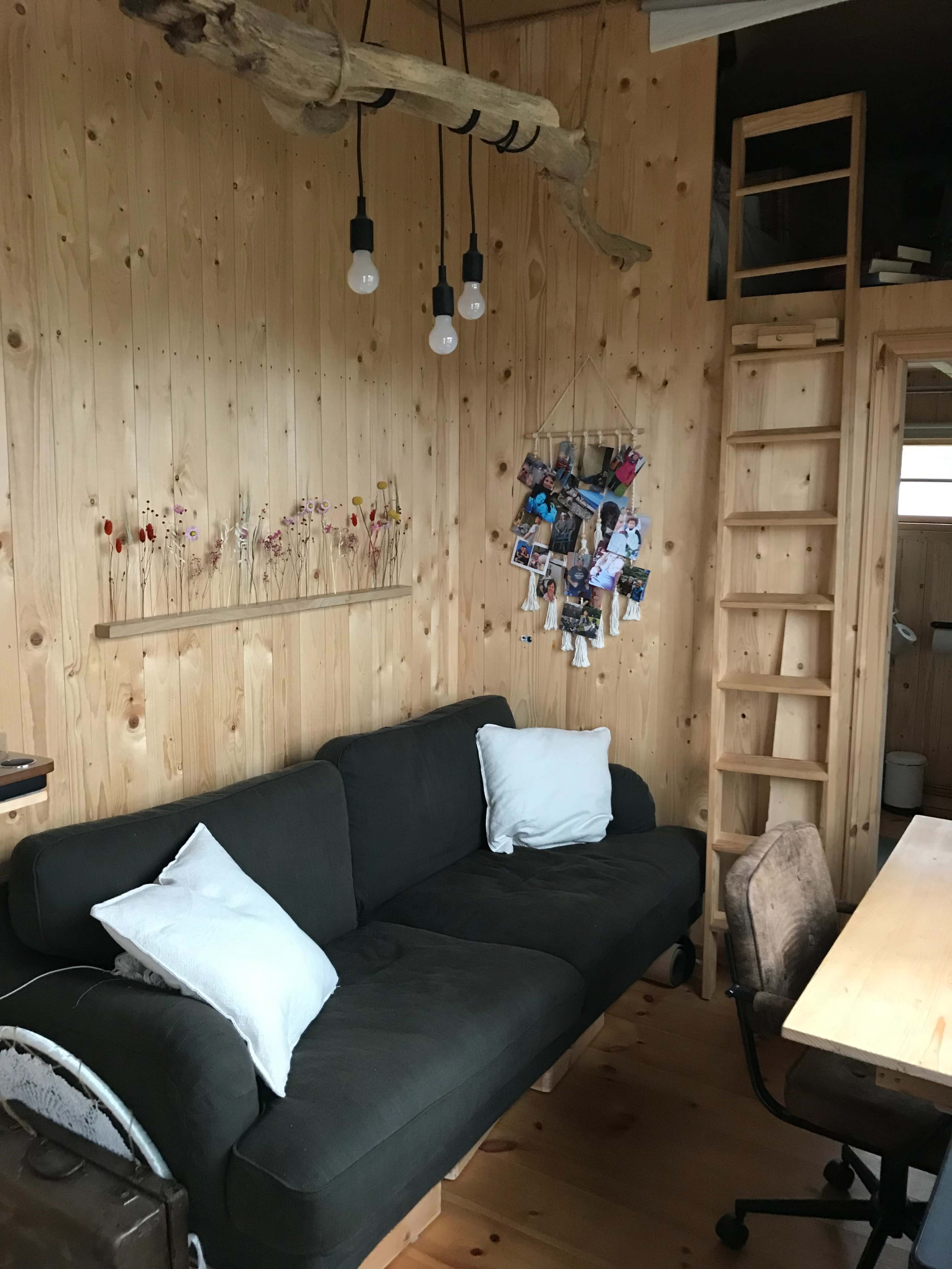 The image shows a cozy room with a wooden interior featuring a dark sofa, a small wooden shelf with flowers, a ladder leading to an upper level, and a desk with a chair.