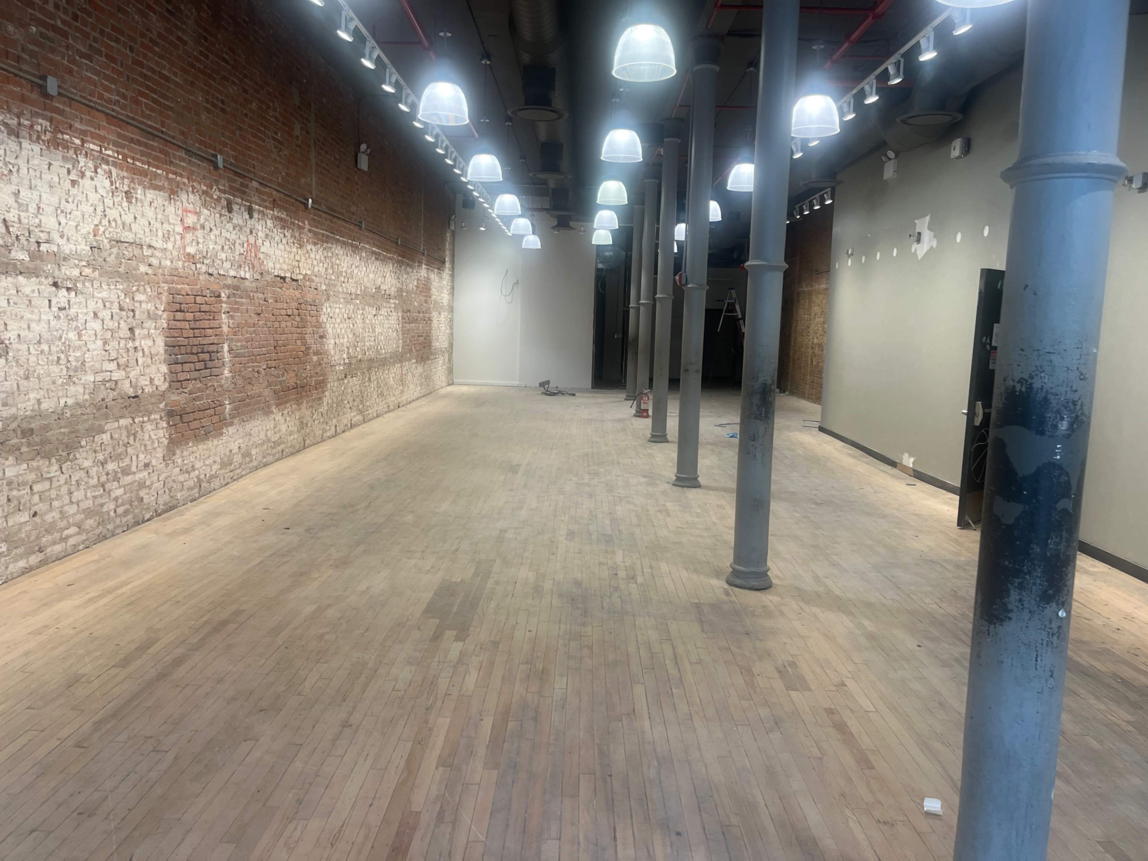 The image shows an empty, industrial-style interior space with exposed brick walls, wooden floors, and several hanging lights.
