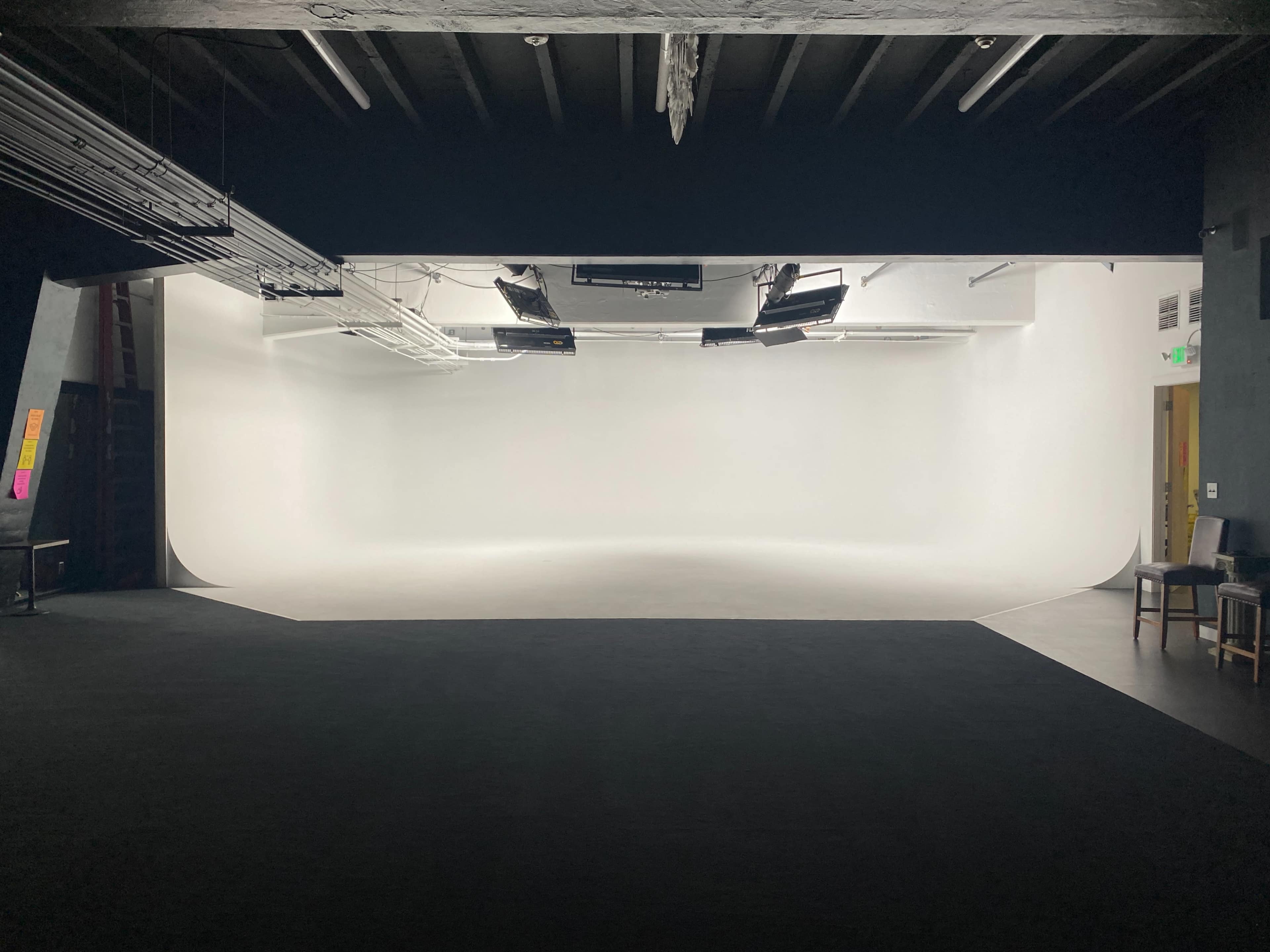 A spacious photography or film studio with a plain white backdrop and dim lighting from overhead fixtures.