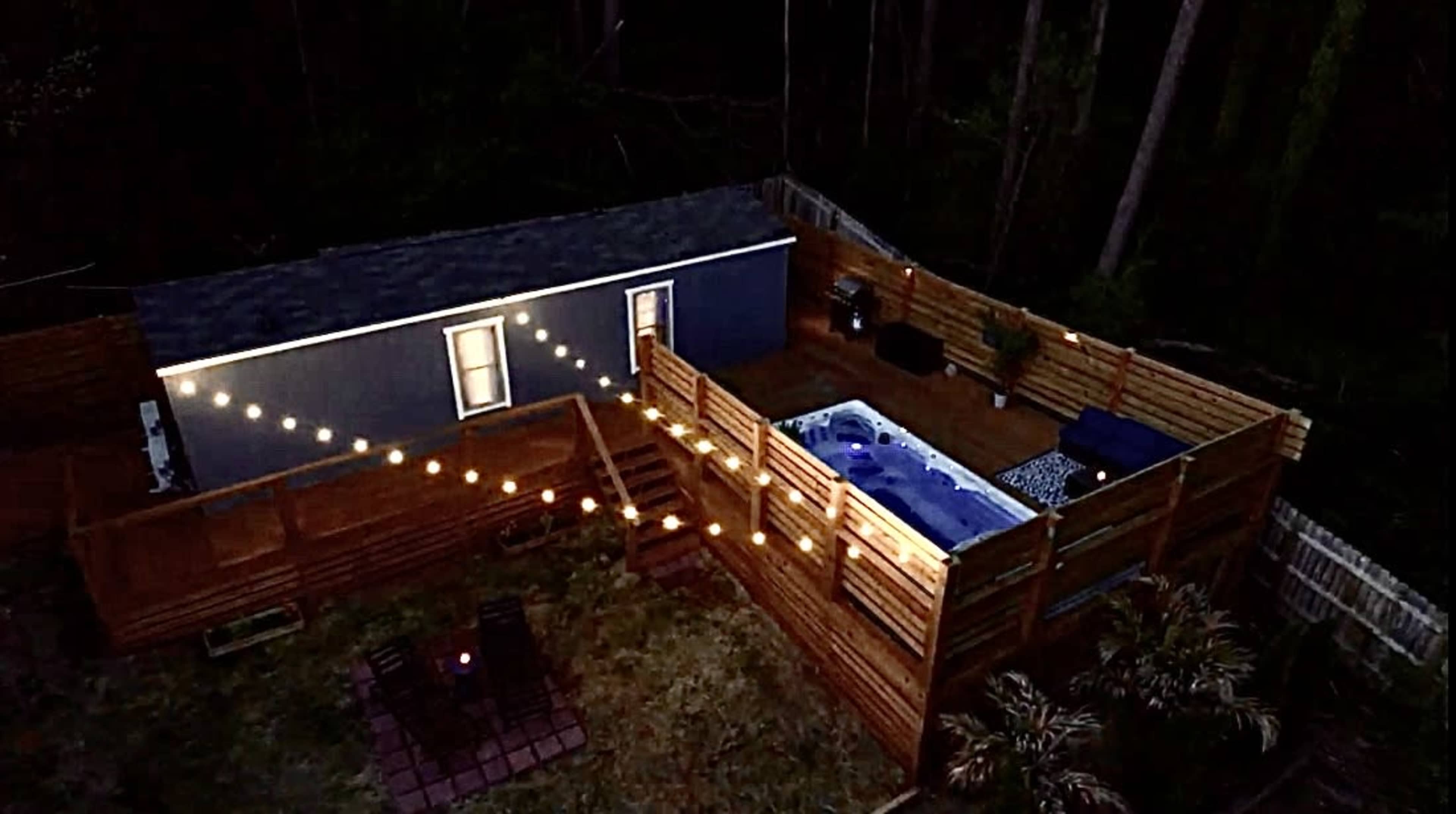 A backyard with a pool surrounded by wooden fencing, a deck, and string lights illuminating the area at dusk.