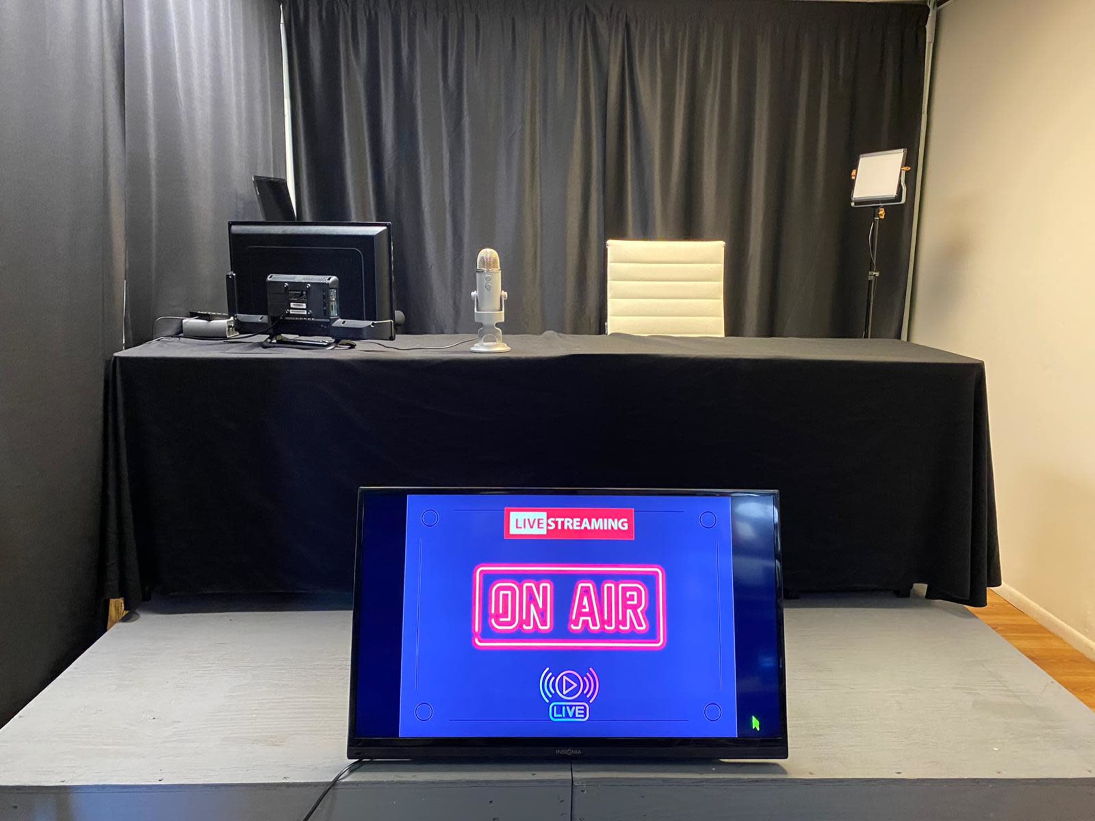 A streaming setup featuring a desk with a microphone, a computer, and a large screen displaying "LIVE STREAMING ON AIR," against a backdrop of black curtains.