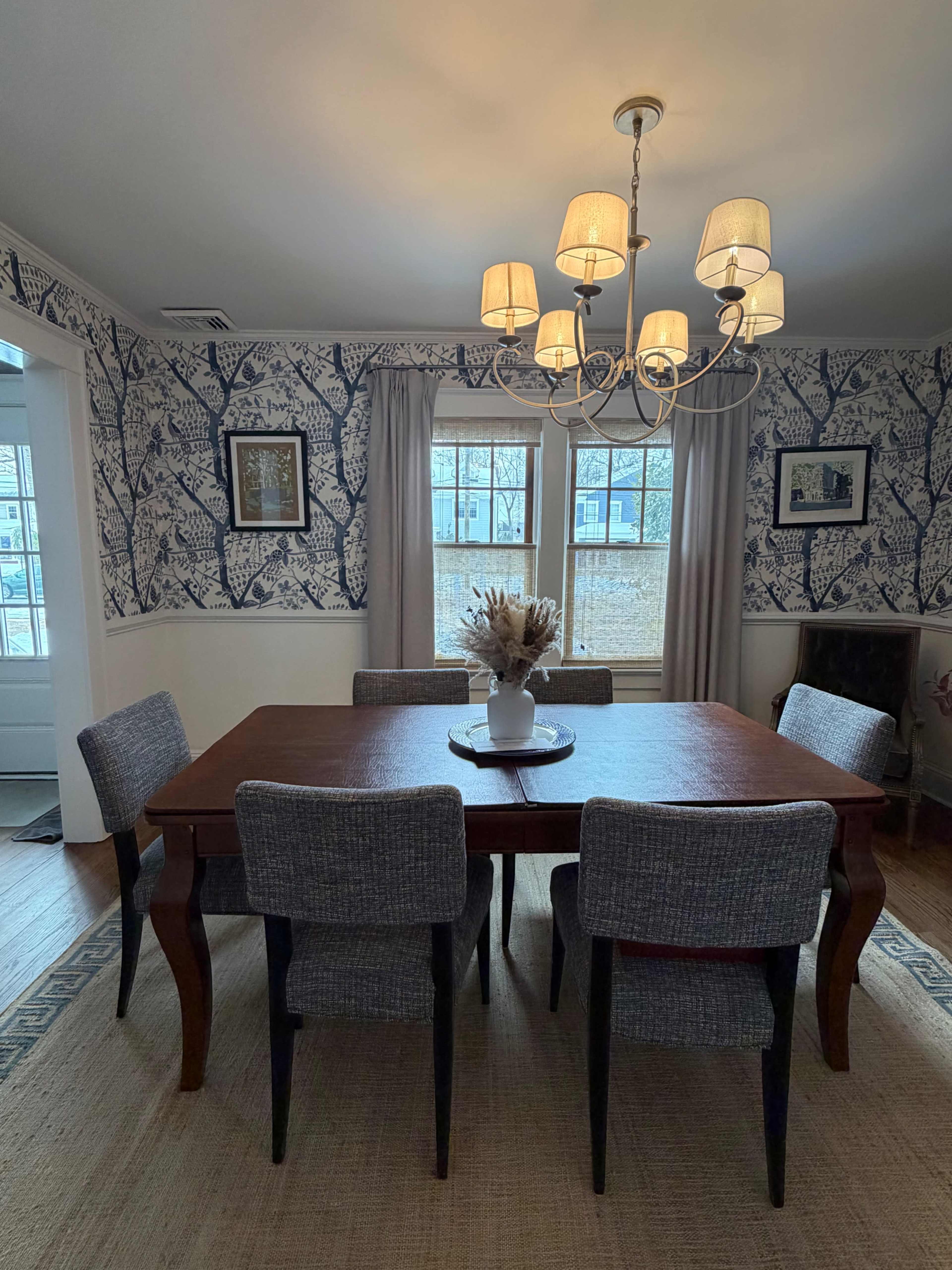 A dining room features a wooden table surrounded by six upholstered chairs, with wallpaper depicting a leaf pattern and a chandelier overhead.