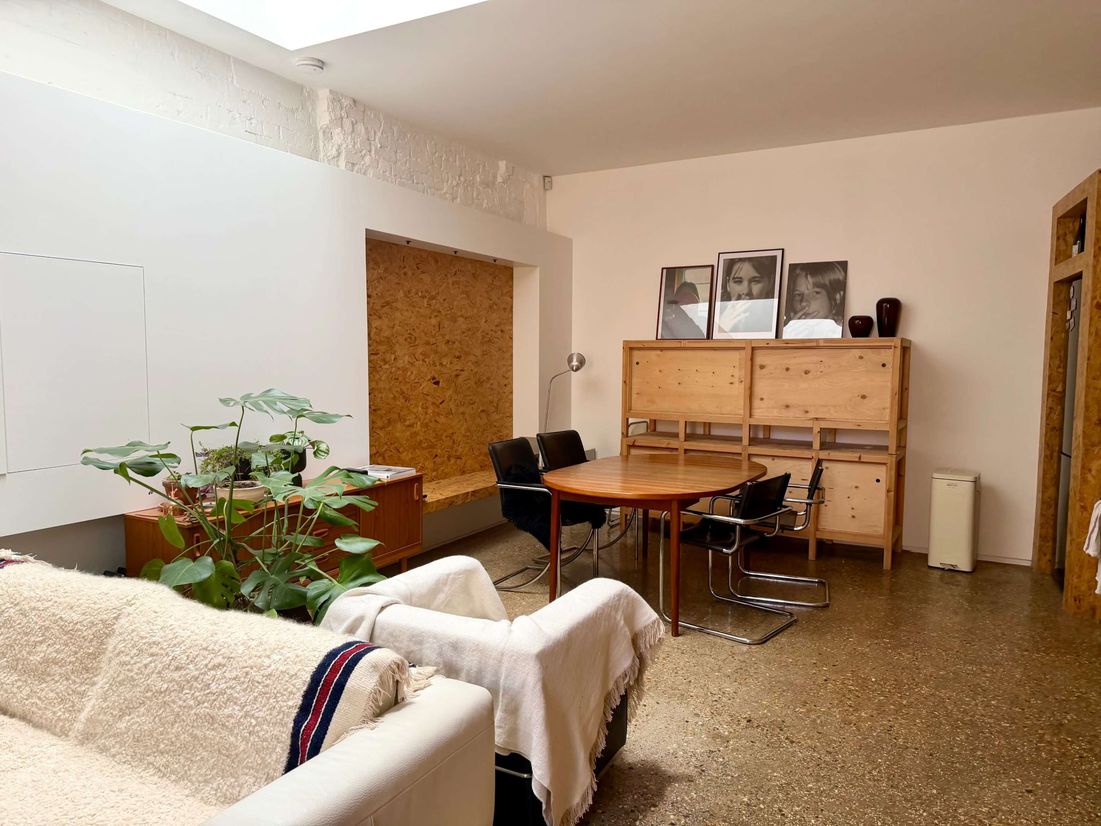 The image shows a modern, minimalist living space featuring a round wooden dining table, black chairs, wooden storage units, and framed photographs on the wall, alongside a green plant and a white couch.