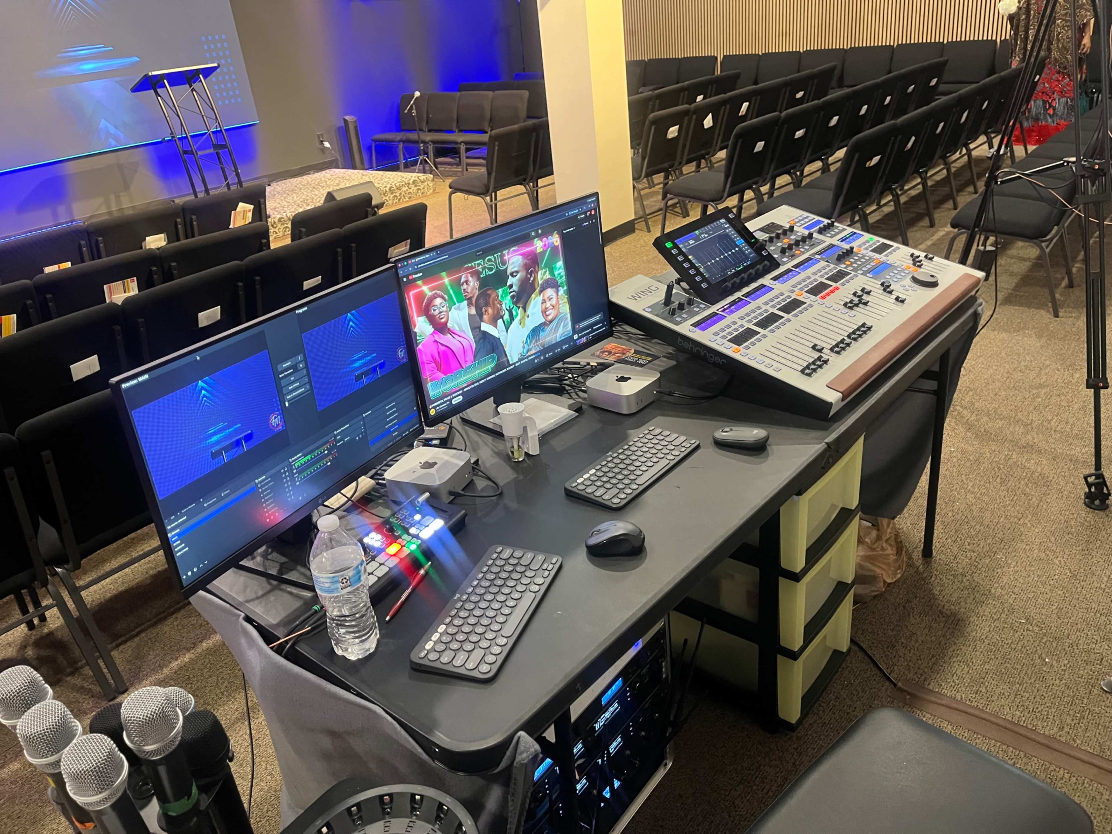 The image shows a control station with multiple screens and a control panel set up in a dimly lit room filled with chairs.