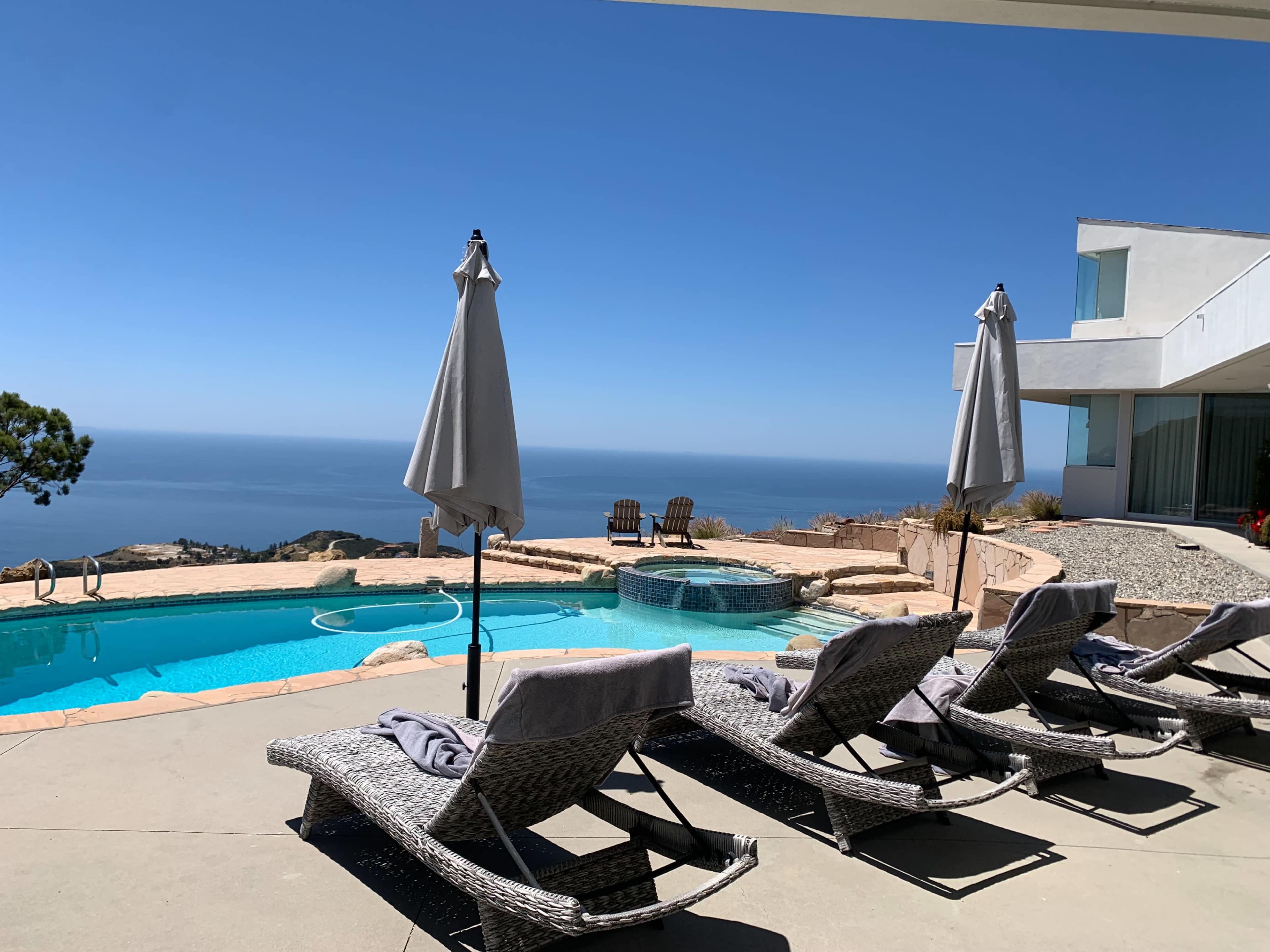 A row of lounge chairs with gray cushions faces a swimming pool overlooking the ocean, with two umbrellas providing shade.