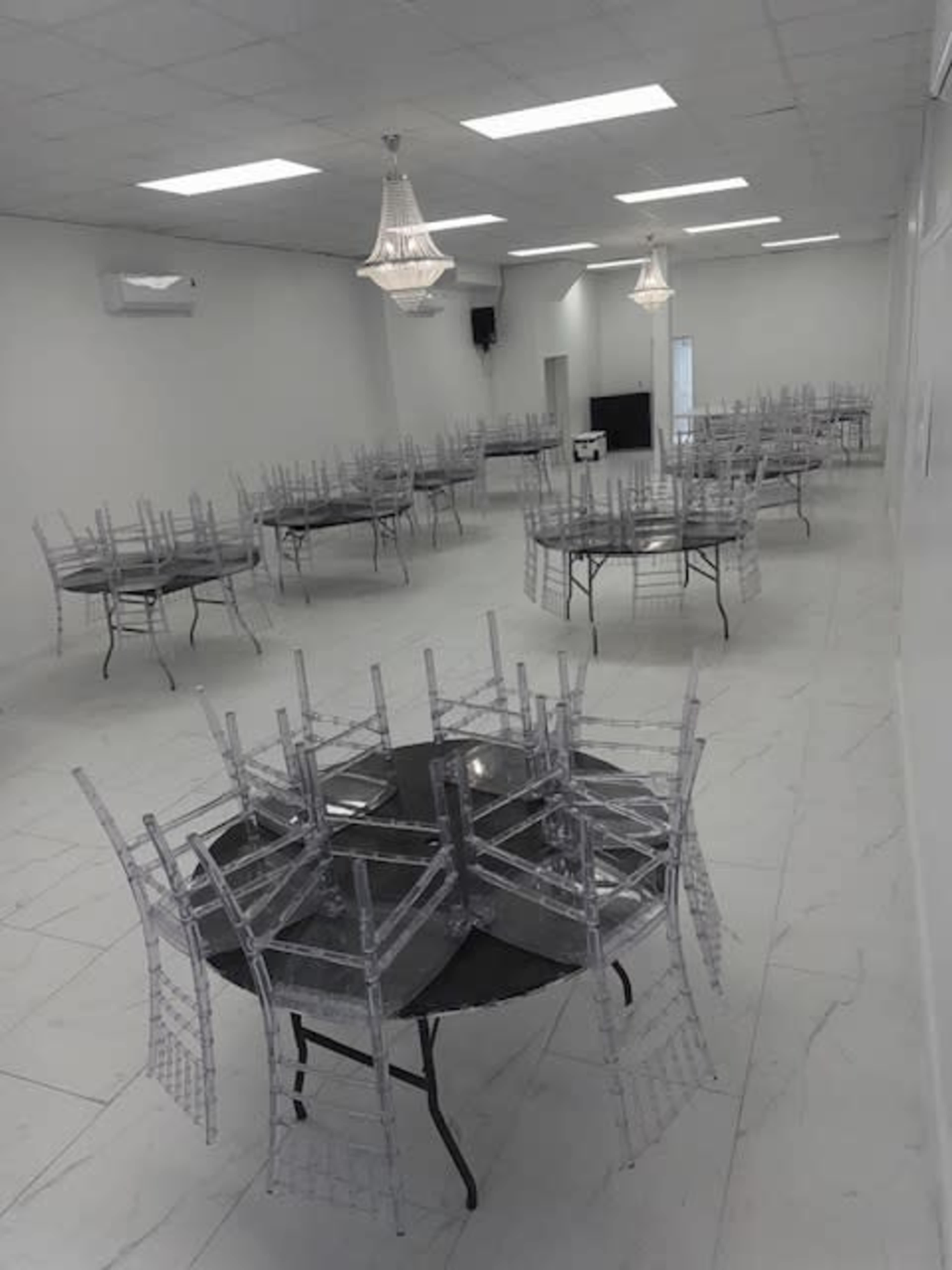 The image shows a spacious, empty room with multiple round tables covered with clear chairs stacked on top, illuminated by overhead lights and featuring a minimalist design.