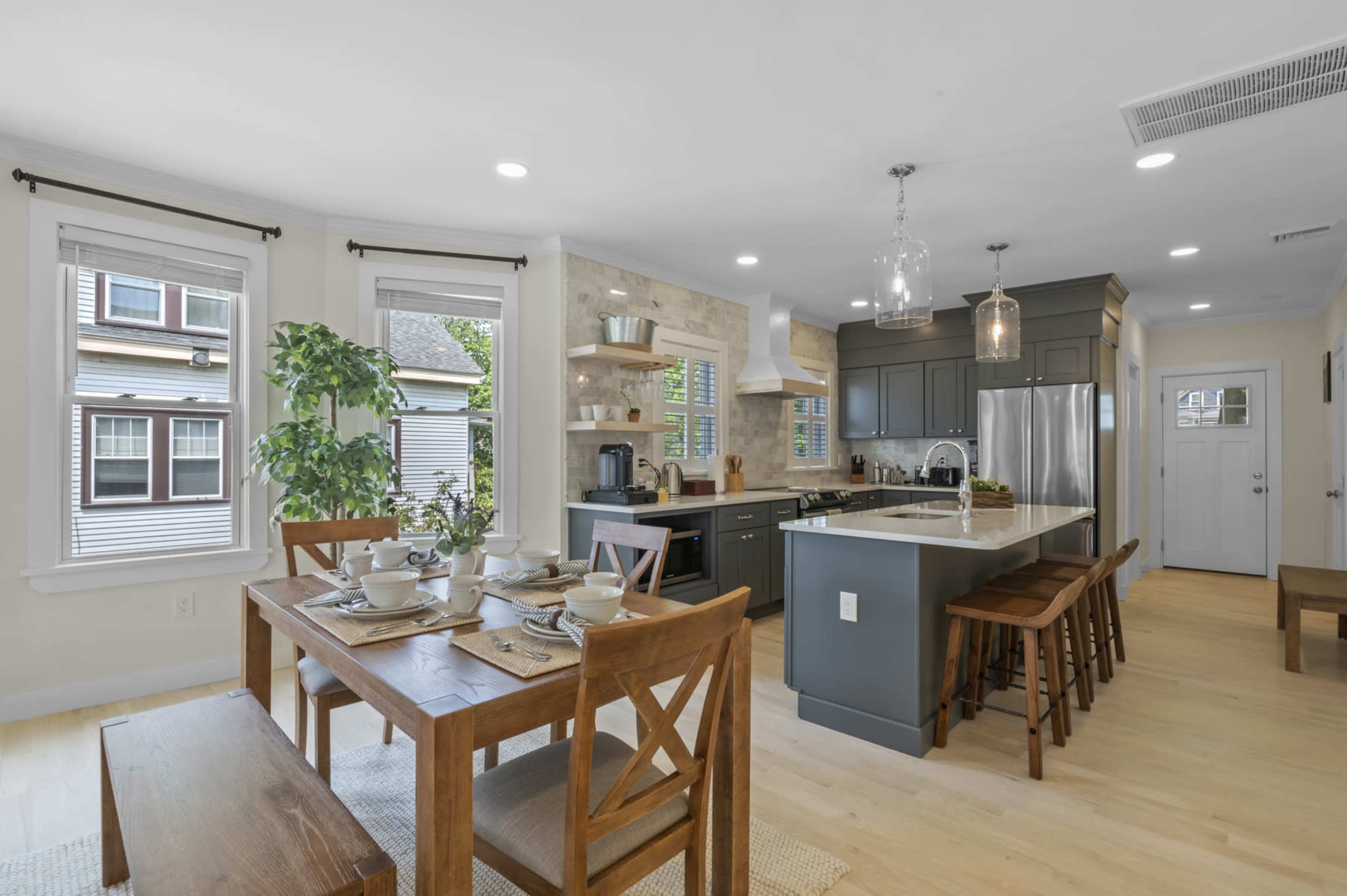A modern kitchen features a central island with seating, a dining table set for four, and large windows allowing natural light to fill the space.