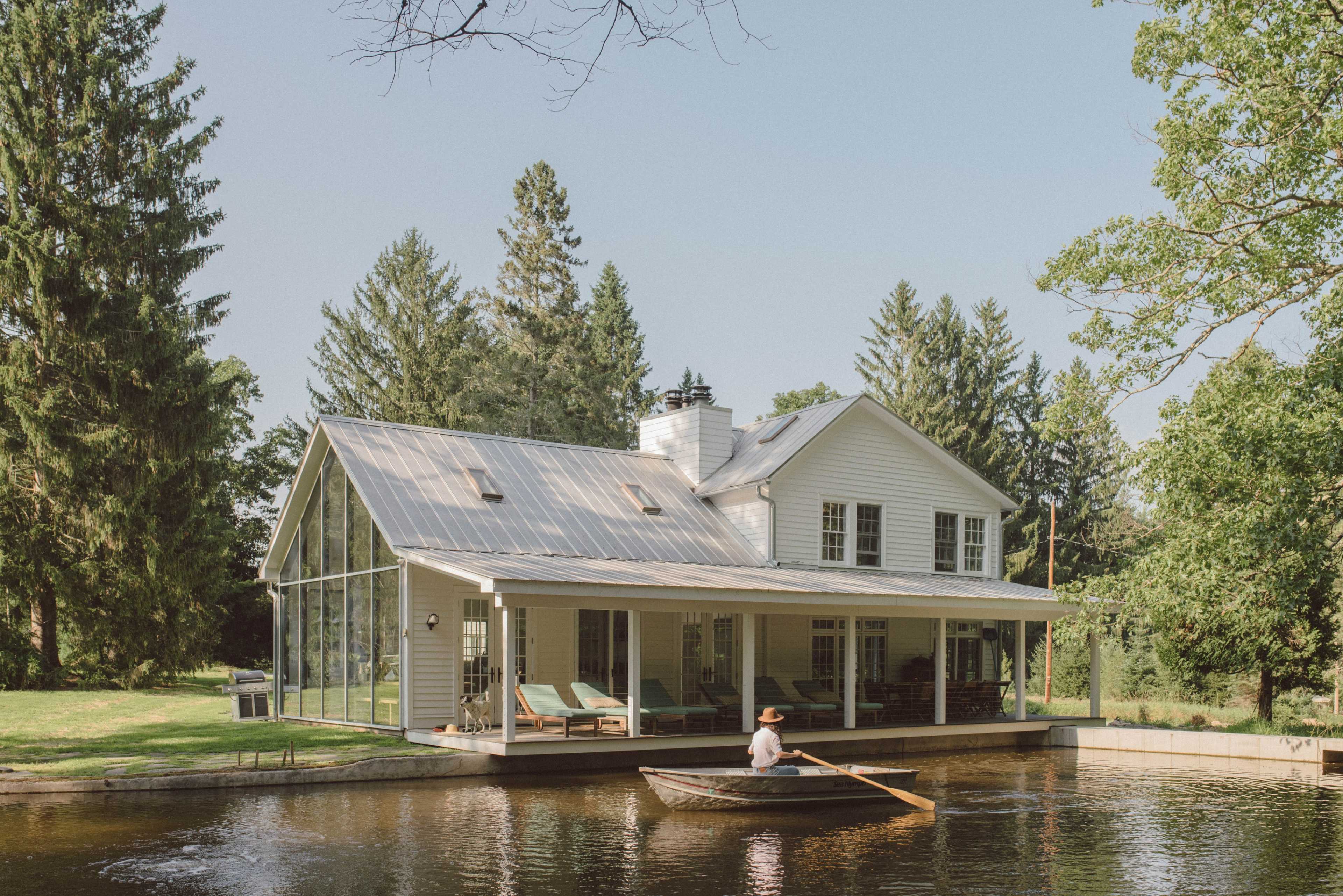 A person rows a small boat on a calm body of water in front of a modern, glass-walled house surrounded by trees.