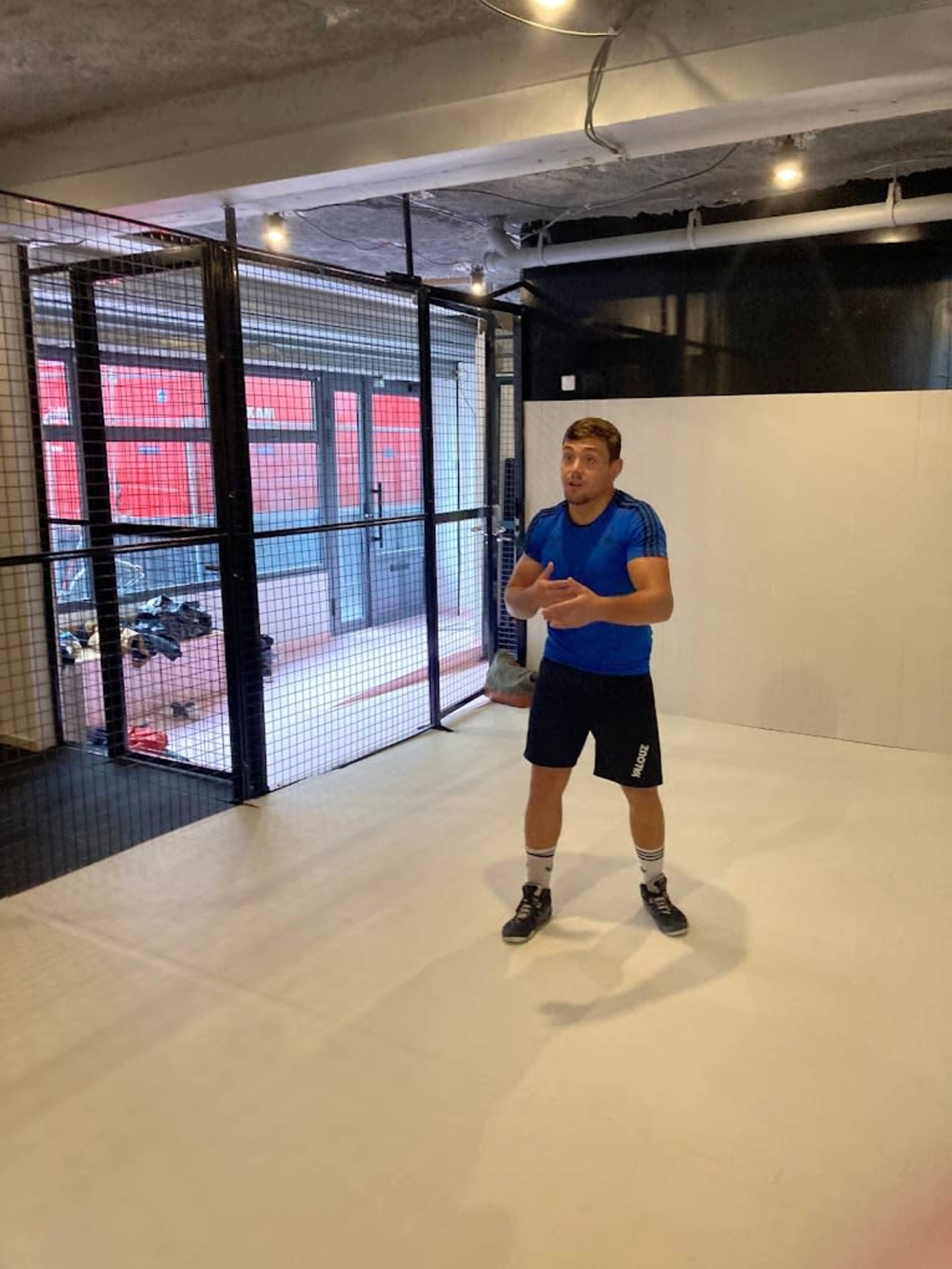A man in a blue shirt and black shorts stands in a well-lit indoor training area surrounded by a netted enclosure.