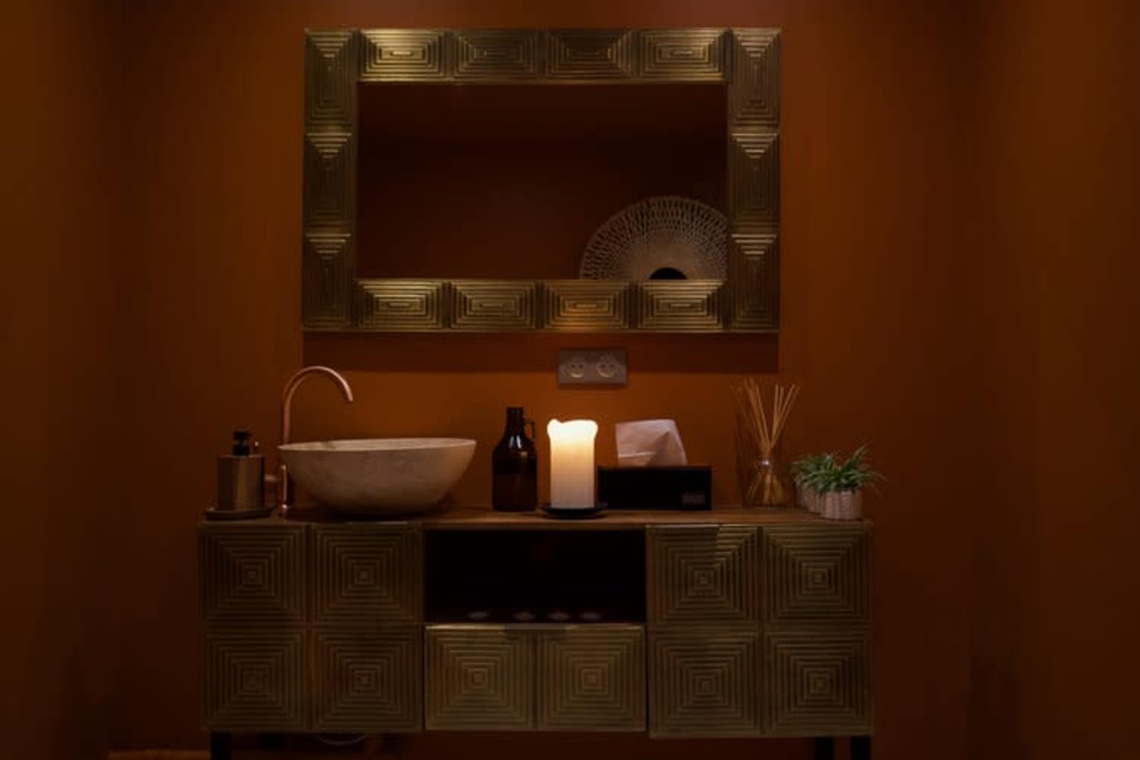 The image shows a well-decorated bathroom vanity with a bowl sink, a lit candle, and neatly arranged decorative items against an orange wall.