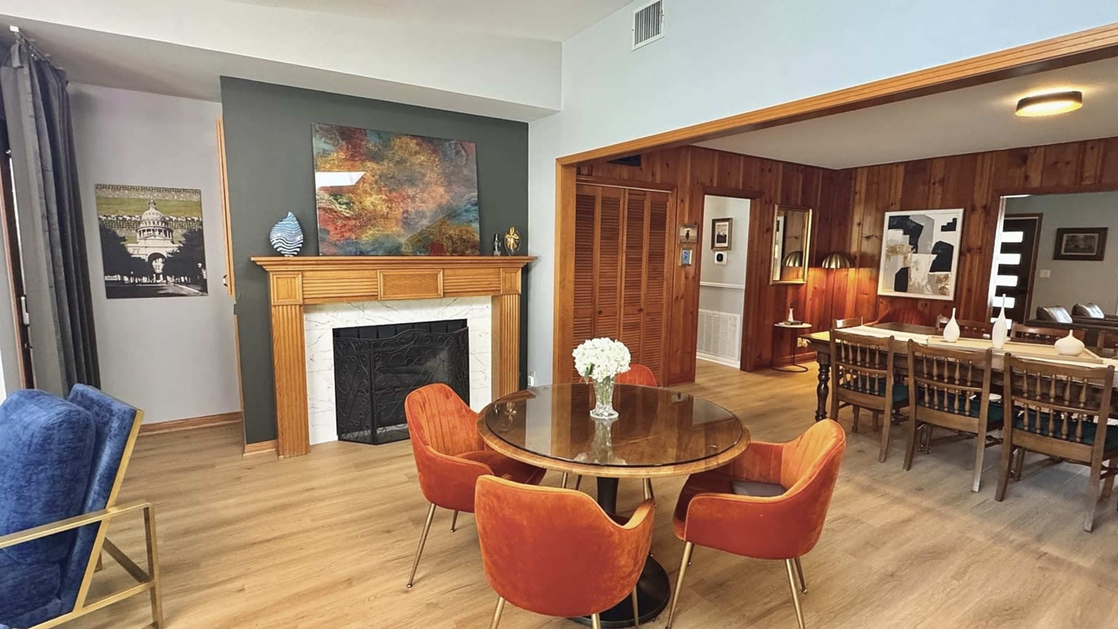 The image shows a living and dining area featuring a round wooden table with orange chairs, a fireplace with a decorative mantle, and art on the walls.