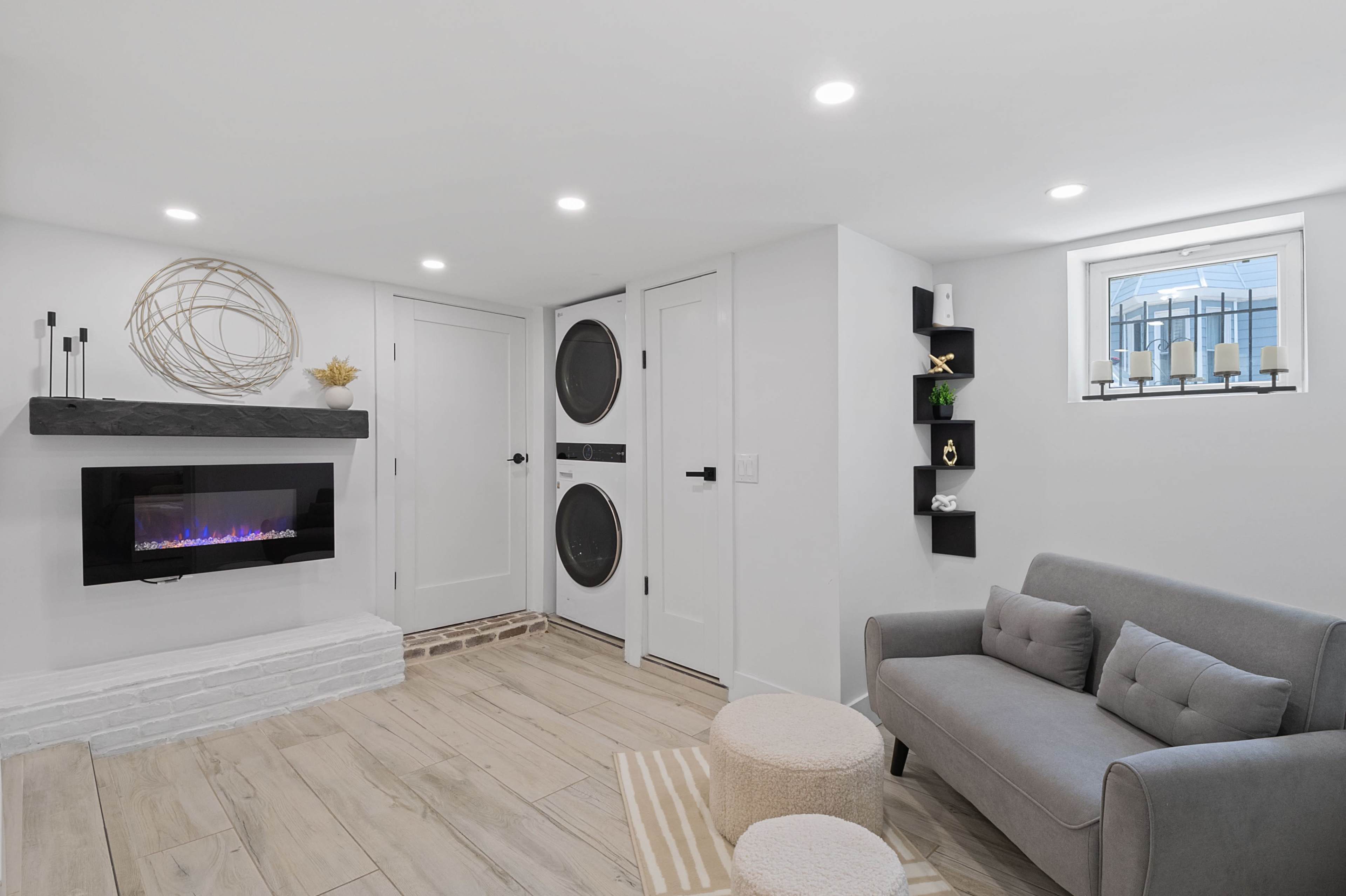 The image shows a modern living space featuring a gray sofa, a small round ottoman, a decorative wall shelf, and a fireplace below a geometric wall decoration, with a washer and dryer unit behind double doors.