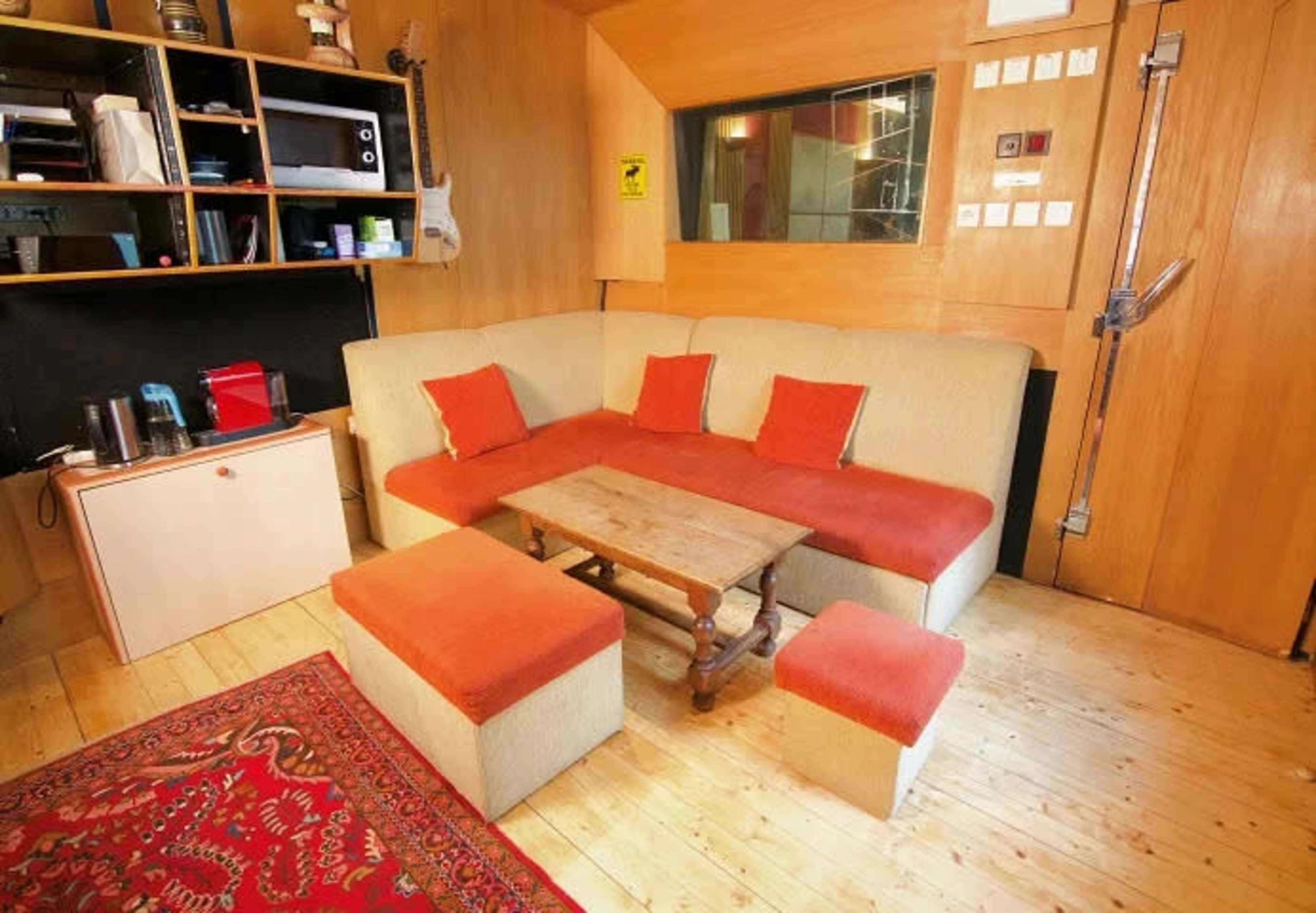 The image shows a wooden-paneled room featuring a couch with orange cushions, a small wooden table, and a shelf with various items, including a guitar and appliances.