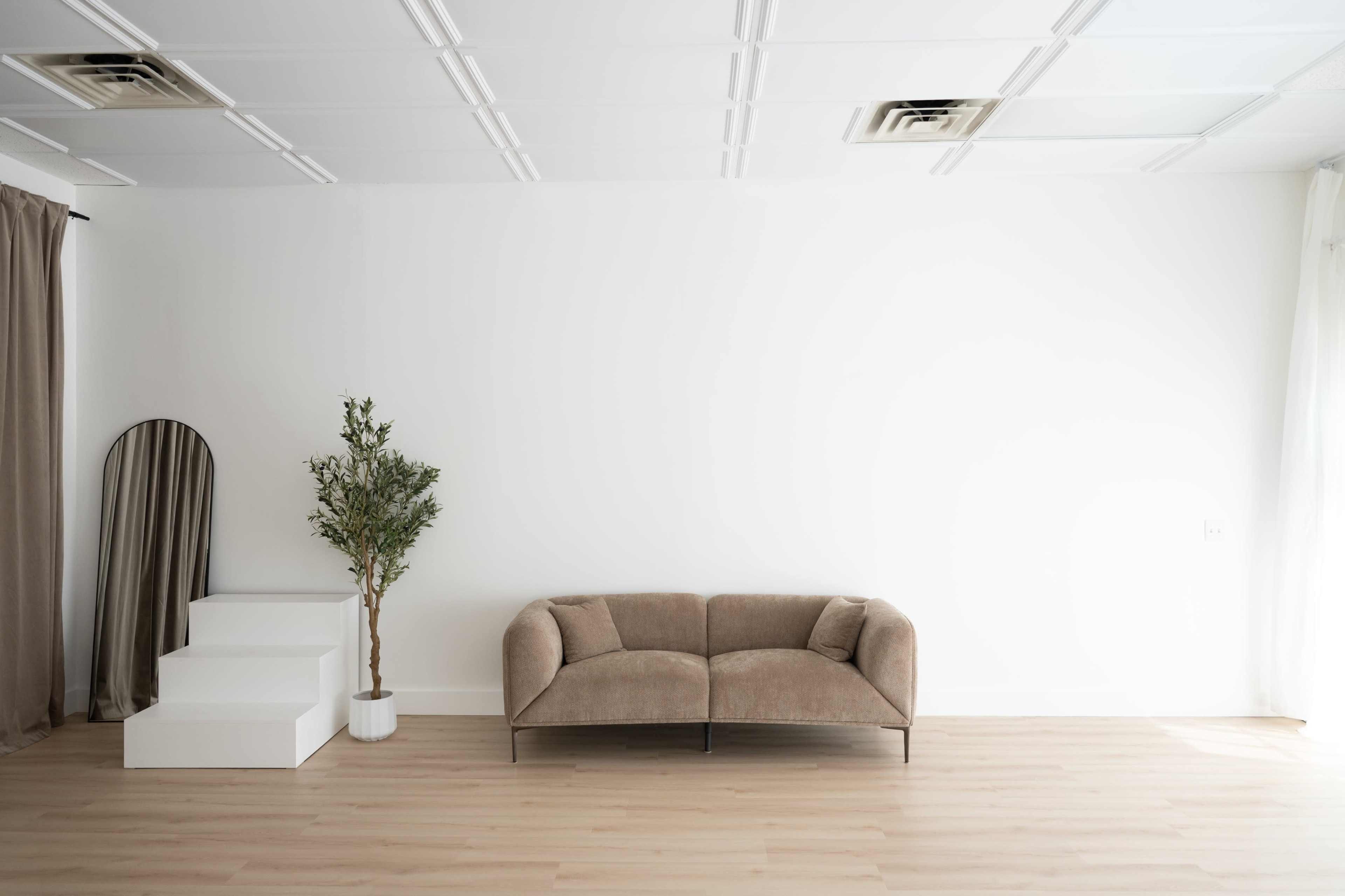 The image shows a minimalist living space featuring a beige couch, a small potted tree, a white staircase, and a large mirror against a plain white wall.