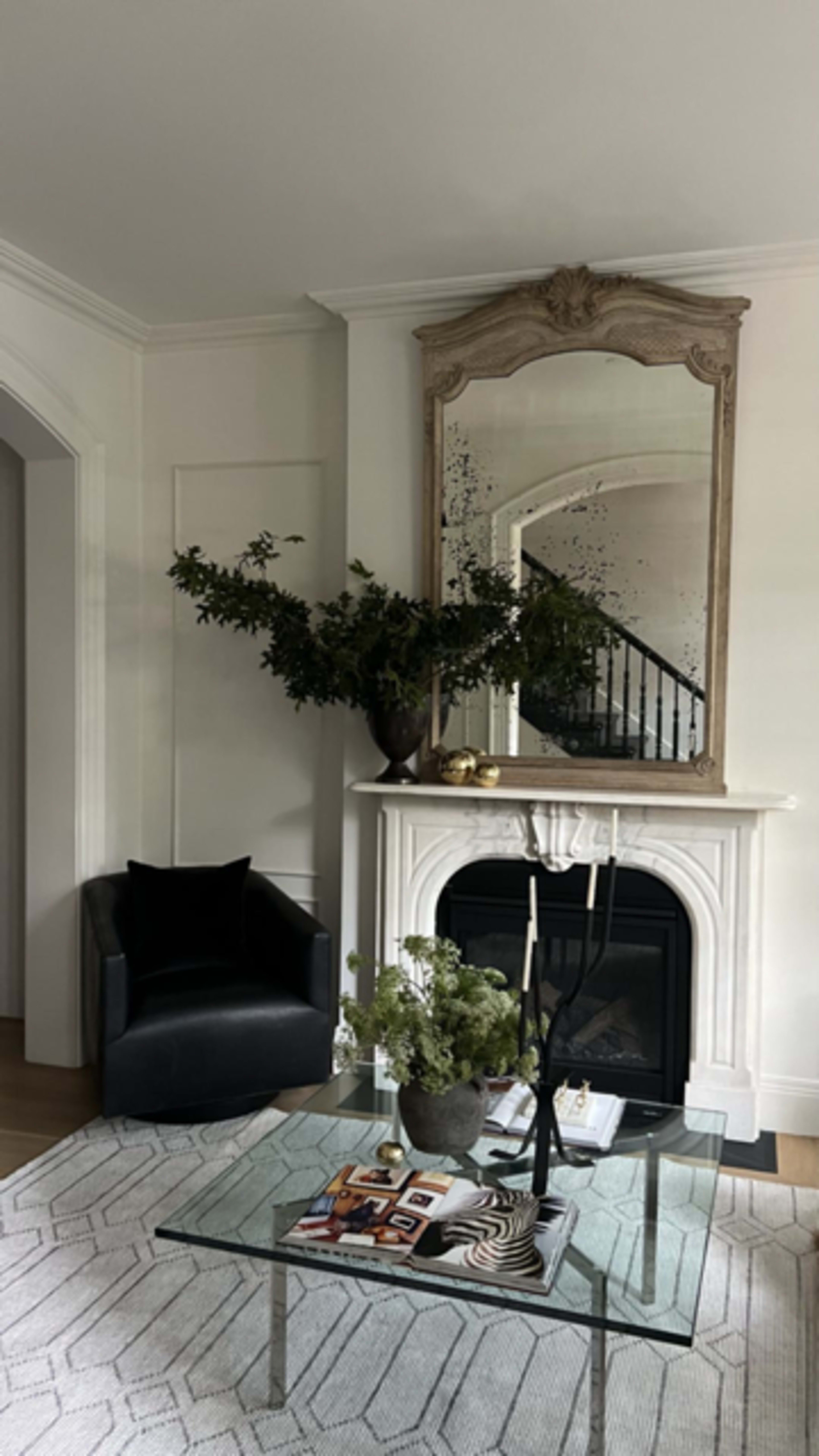 A modern living room features a black armchair, a glass coffee table with decorative items, and a fireplace adorned with a large ornate mirror and a vase of greenery.