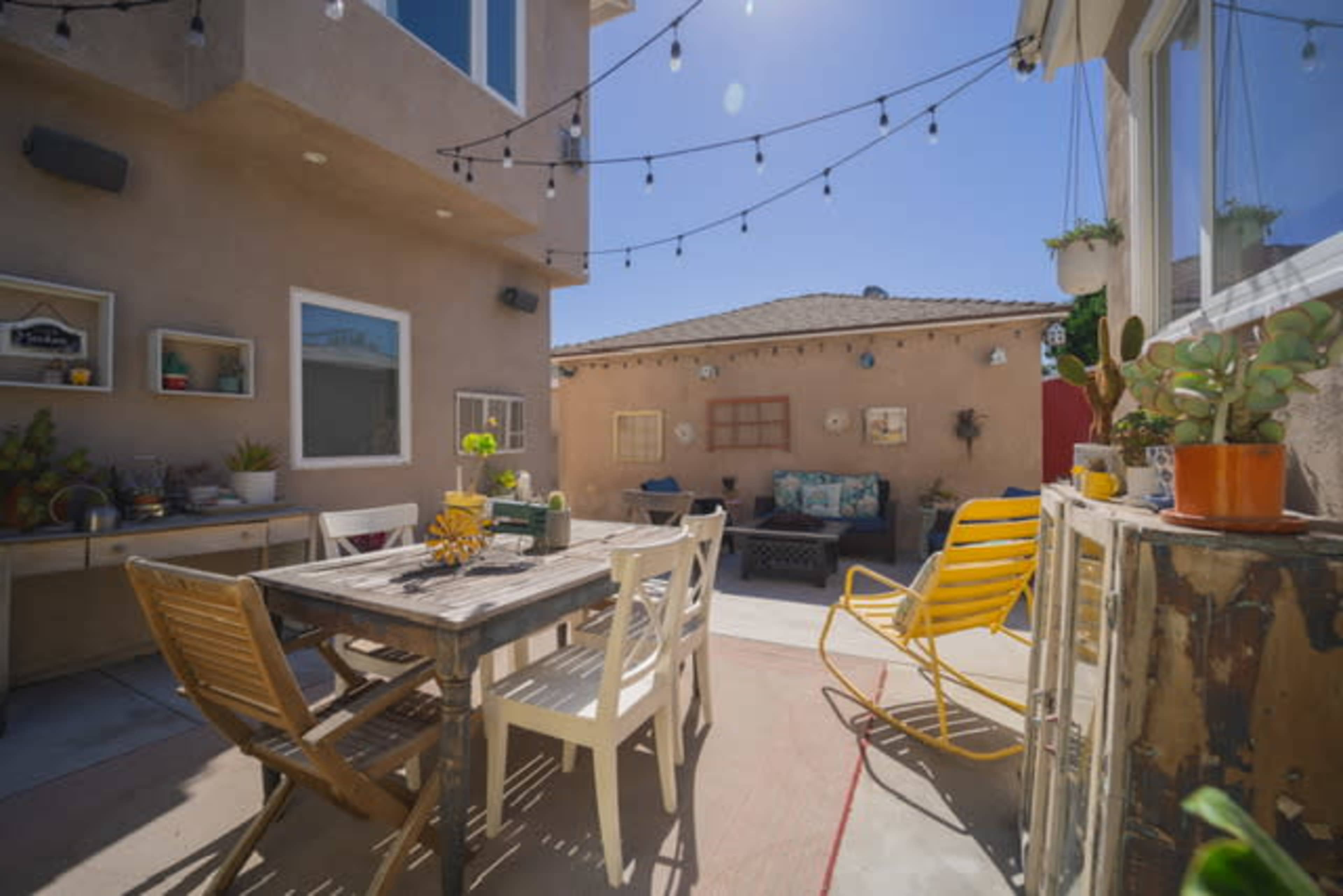 A sunny outdoor patio with a wooden dining table surrounded by chairs, string lights above, and potted plants along the walls.