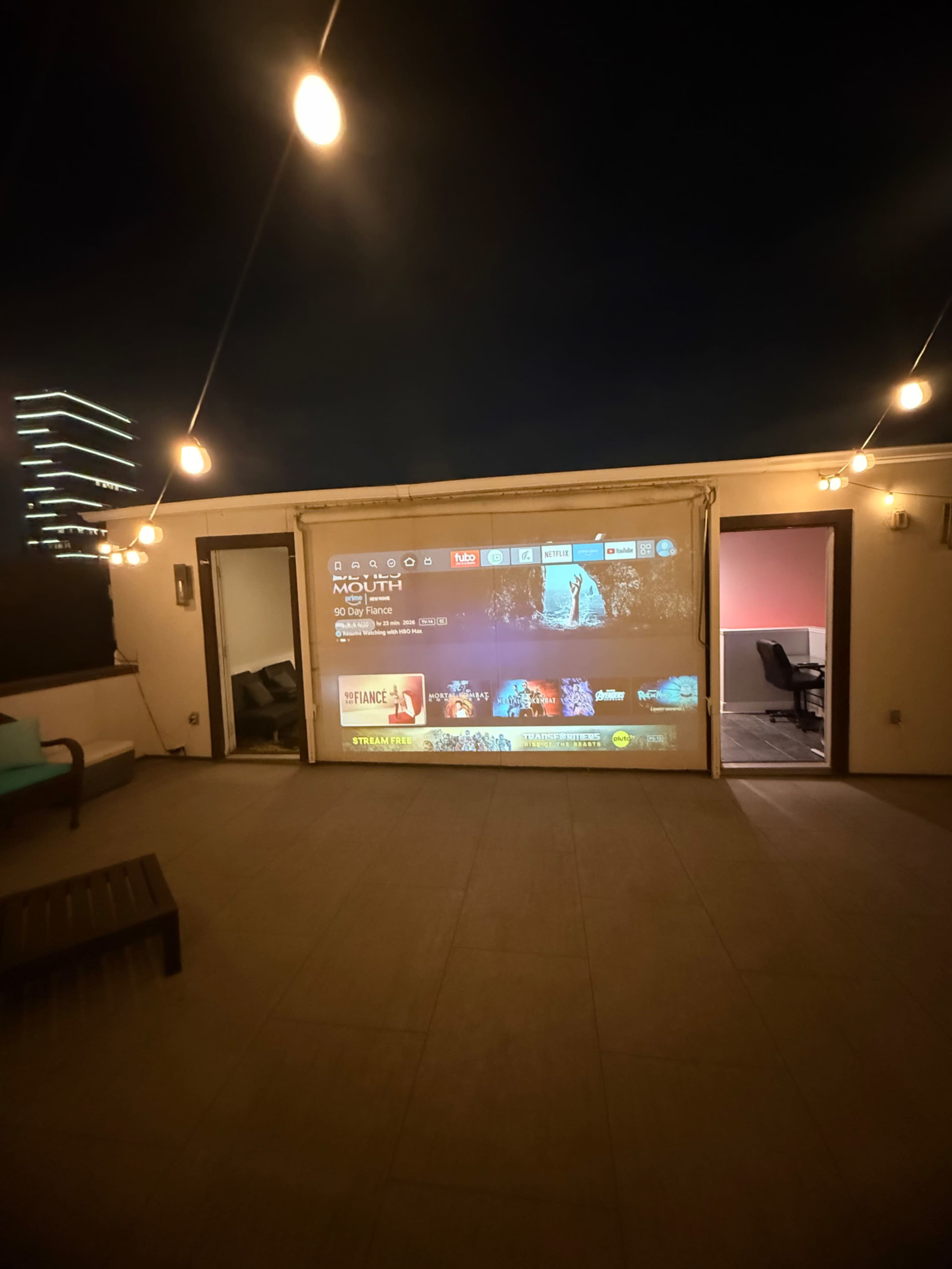 A large outdoor projection screen displays a movie interface on a rooftop, surrounded by string lights and modern furniture.