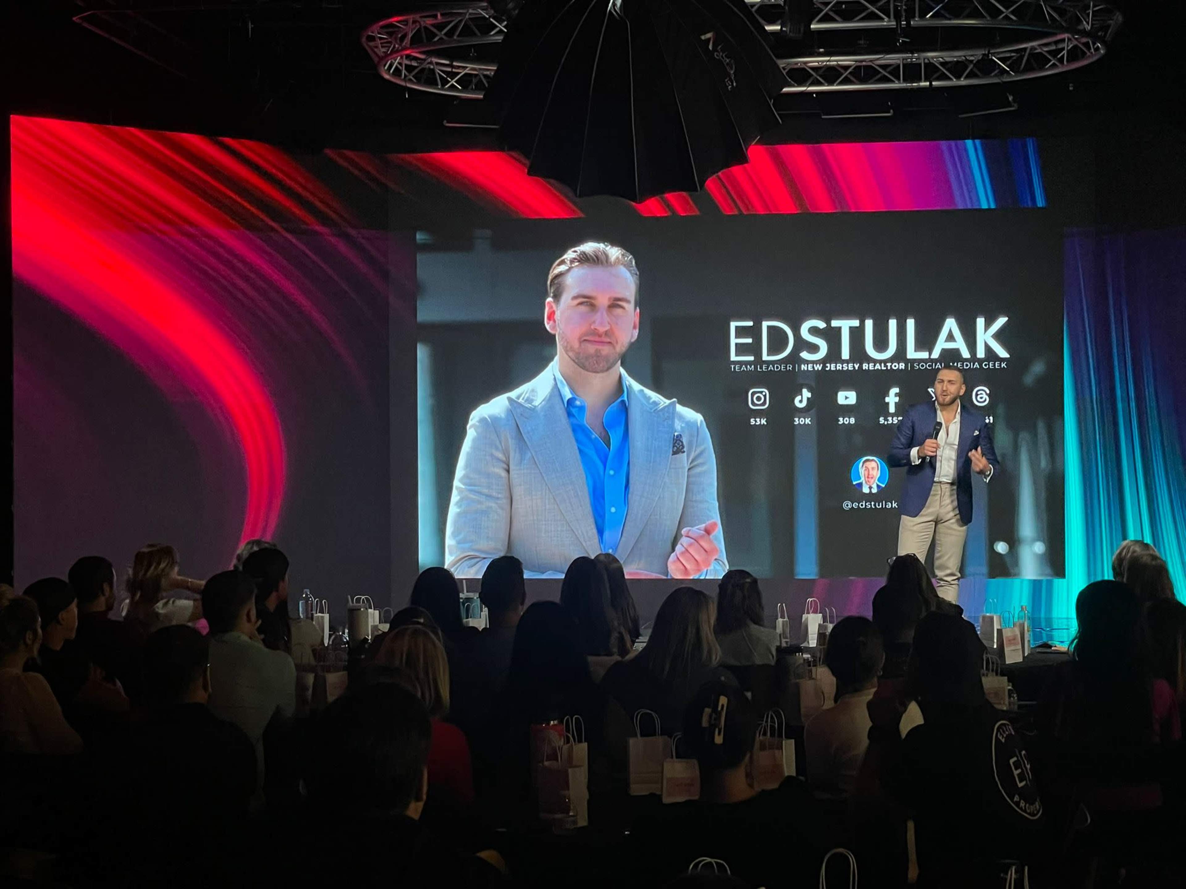 A speaker presents on stage to an audience while a large screen displays a portrait and information about Ed Stulak.