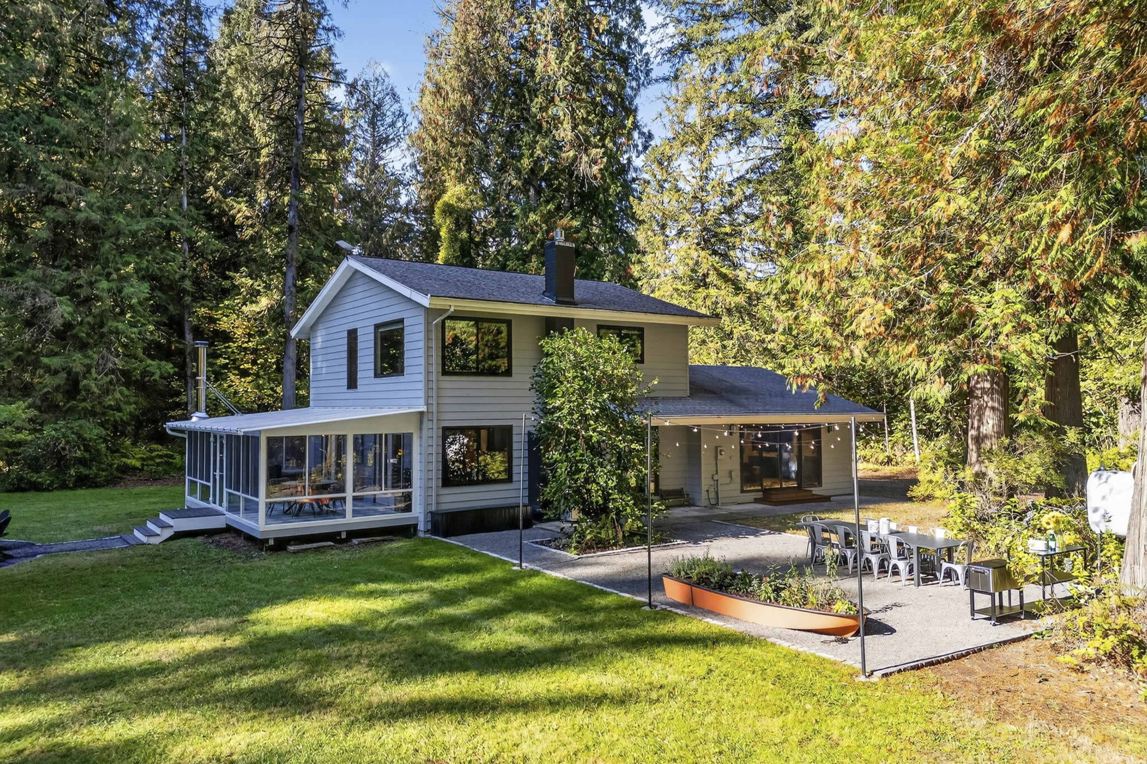 A two-story house with a screened porch and a stone patio is surrounded by tall trees in a grassy area.