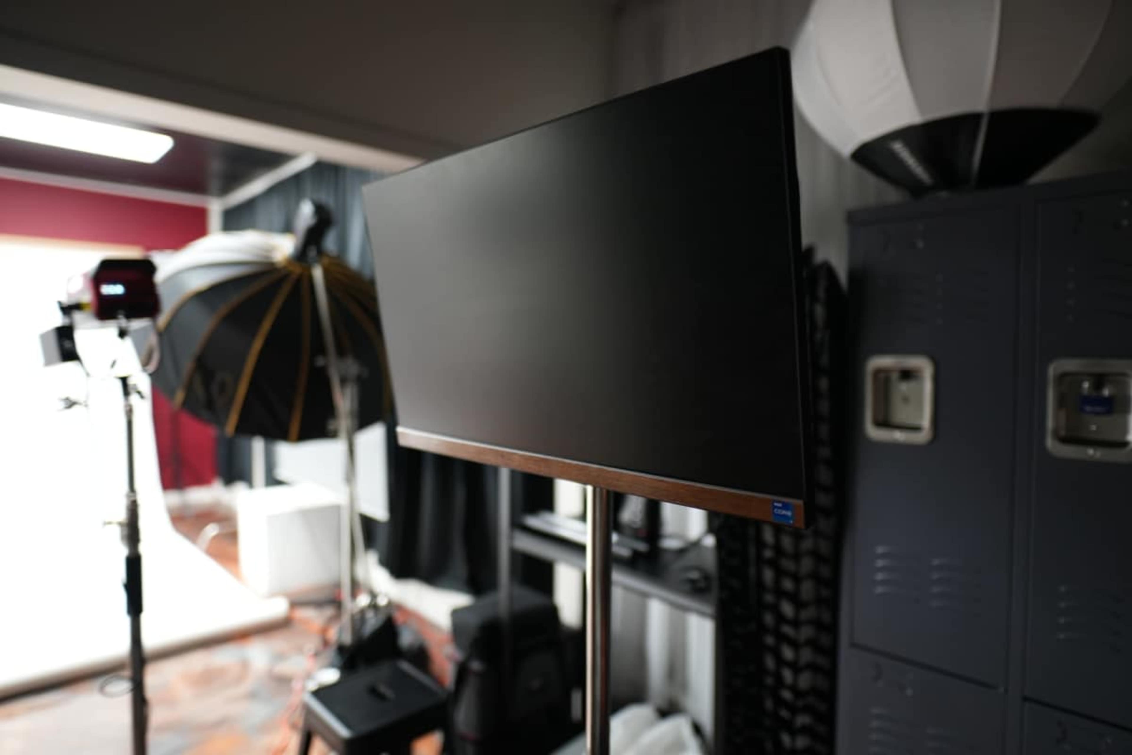A blank monitor is mounted on a stand in a photography studio, surrounded by lighting equipment and storage lockers.