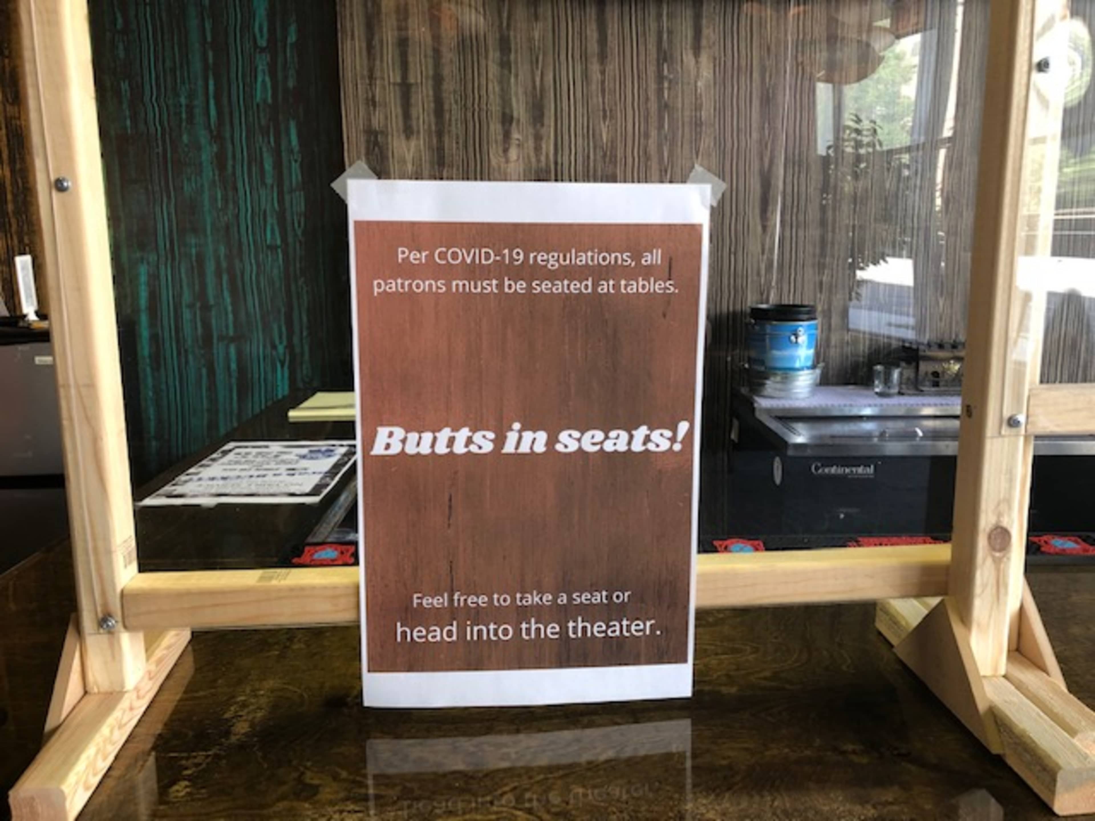 A sign on a wooden surface informs patrons that, per COVID-19 regulations, they must be seated at tables and encourages them to find a seat or head into the theater.