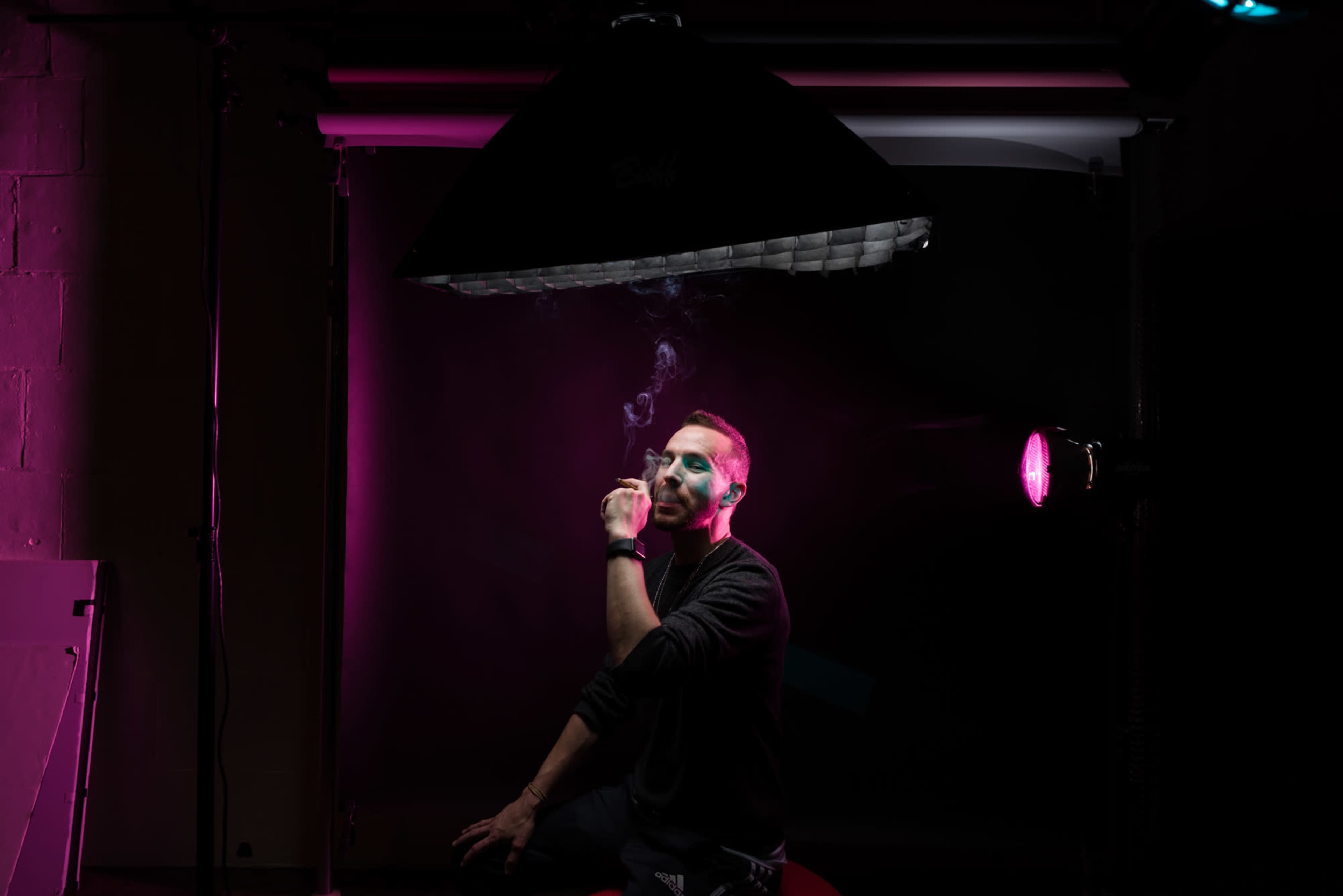 A man sits in a dimly lit studio with pink lighting, holding a cigarette and exhaling smoke.