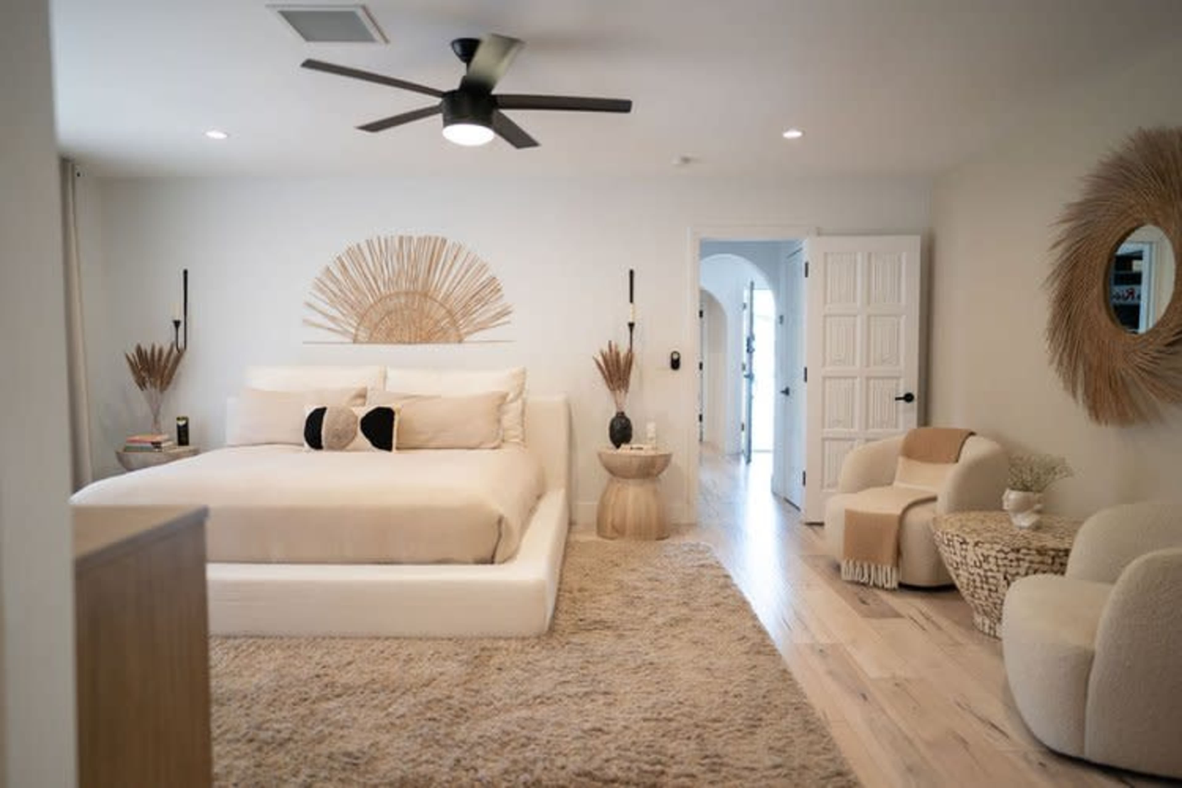 The bedroom features a large bed with neutral bedding, a round mirror, and minimalist furniture, all set on light wood flooring.