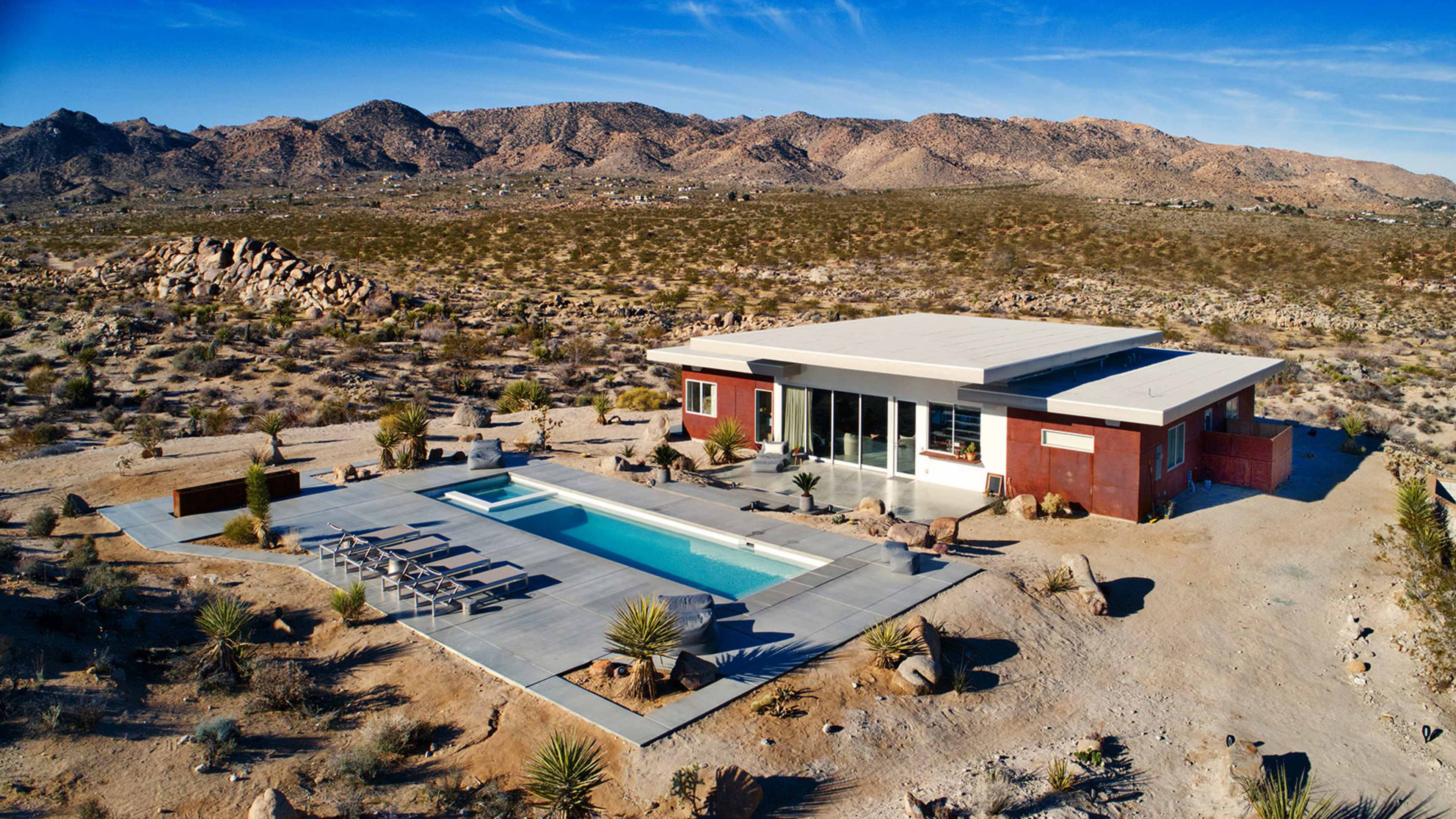 A modern house with a swimming pool is situated in a desert landscape surrounded by mountains and sparse vegetation.