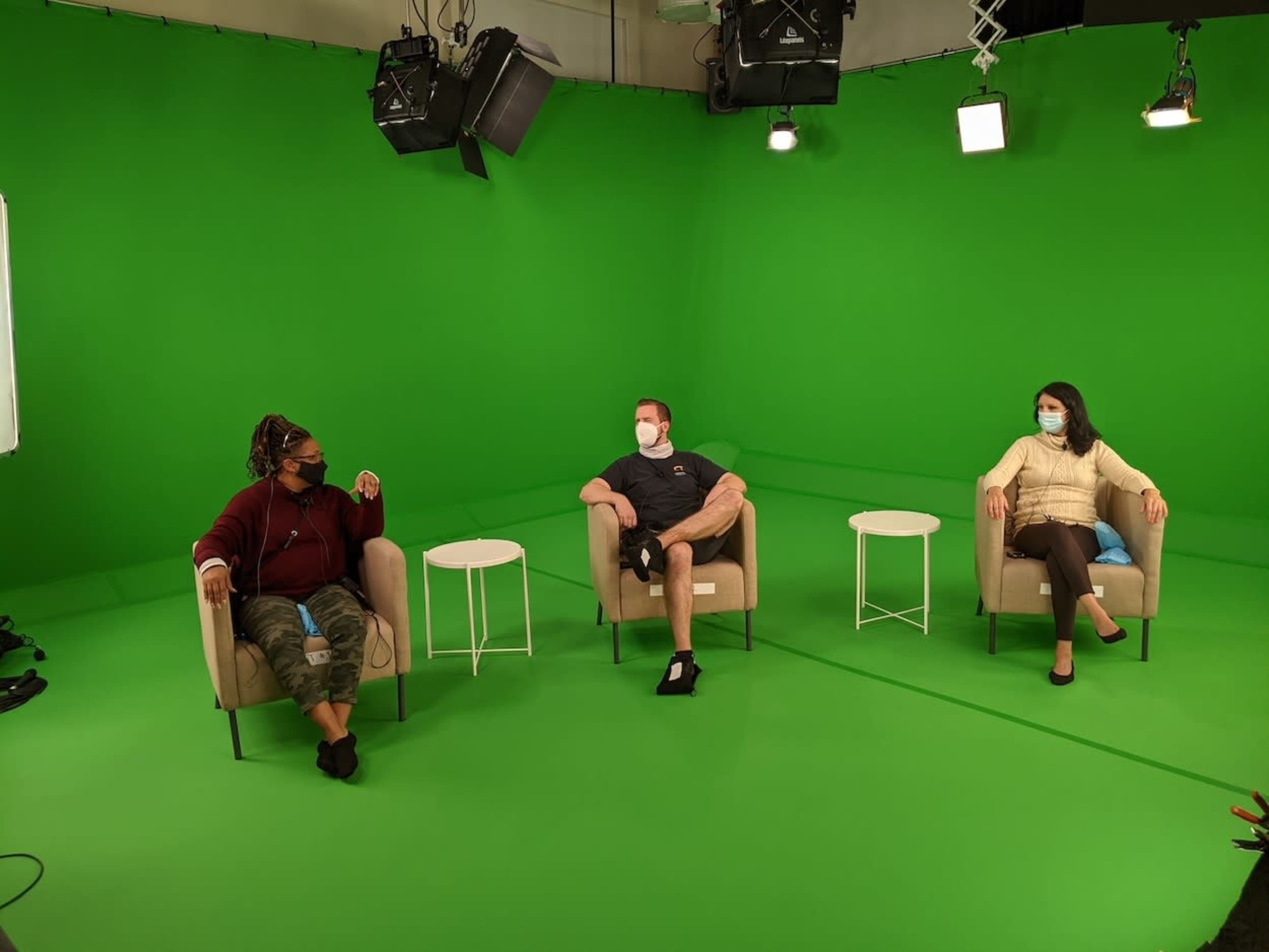 Three individuals sit in a green screen studio, wearing masks, on chairs near small white tables.