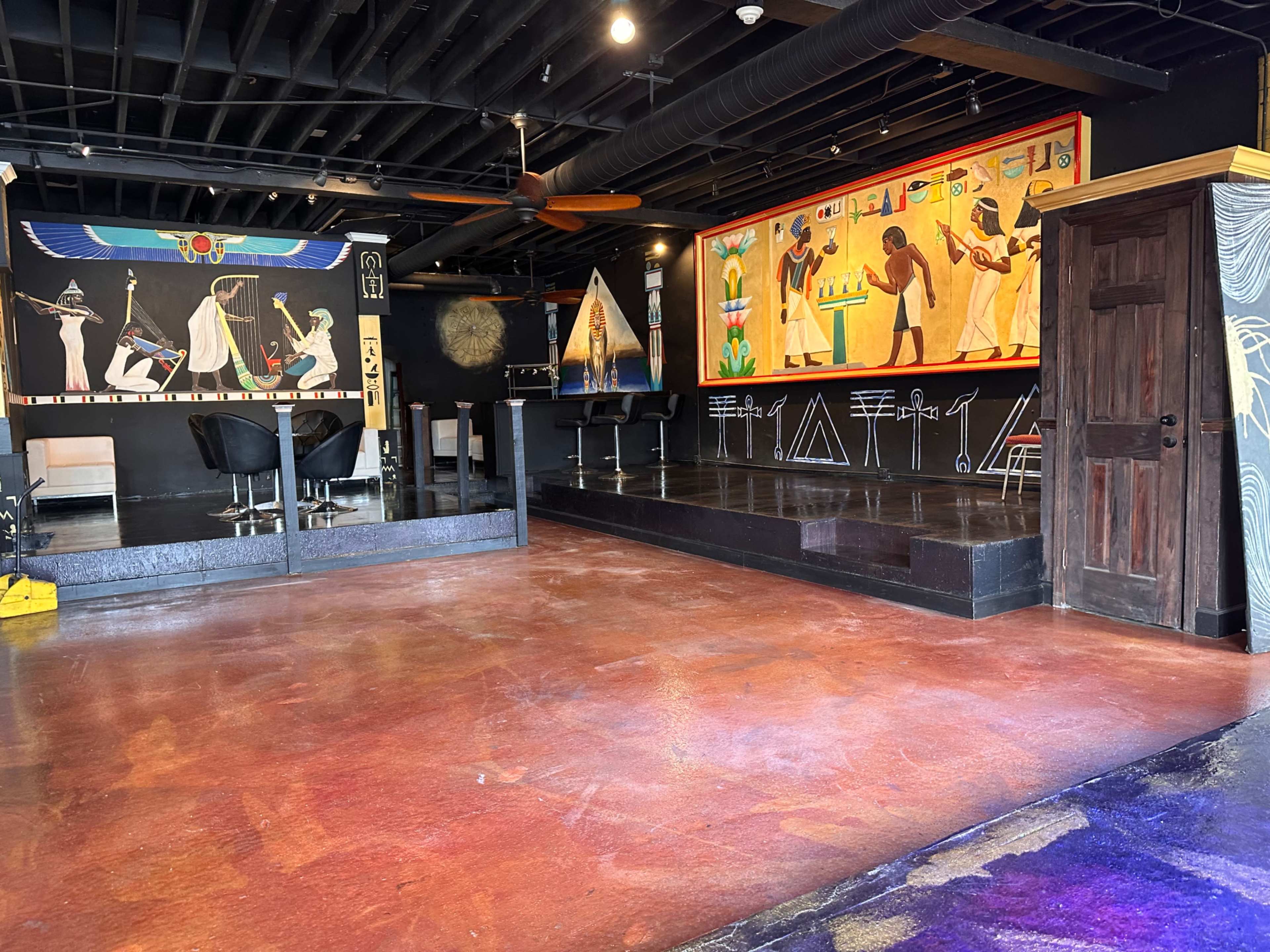 A spacious, dimly lit room with a stage area, decorated with ancient Egyptian mural artwork on the walls and a bar counter along one side.