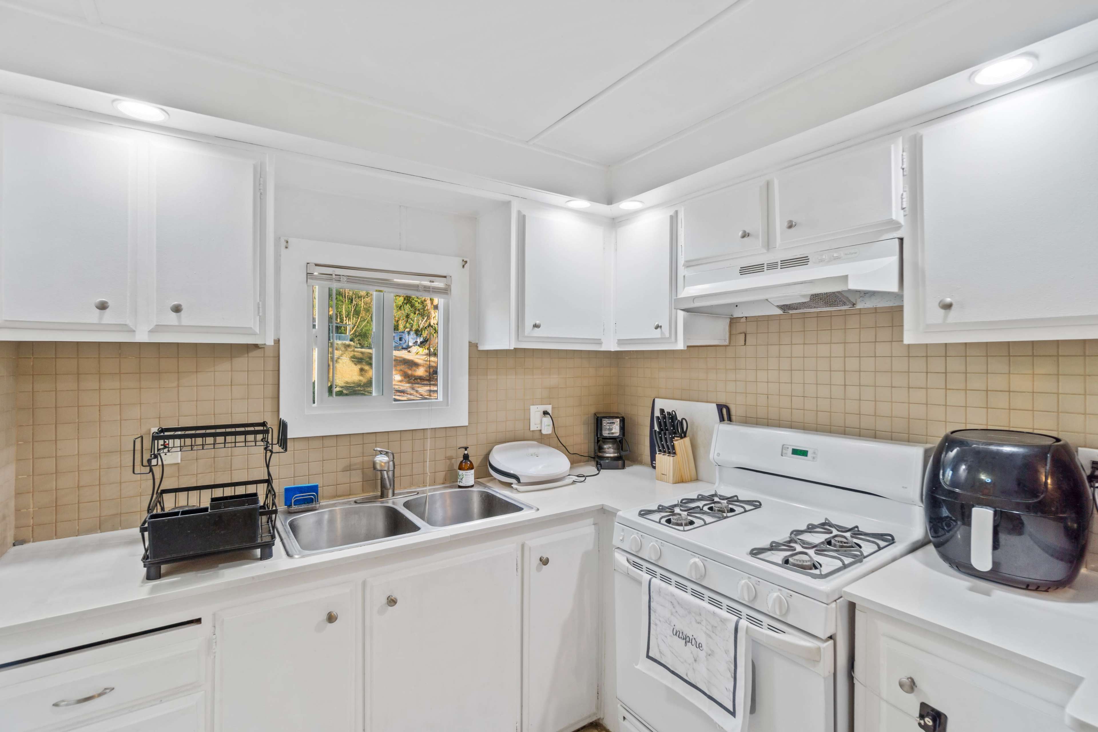 A modern kitchen features white cabinets, a double sink, a stove with an oven, and plumbing, with a window allowing natural light to enter.